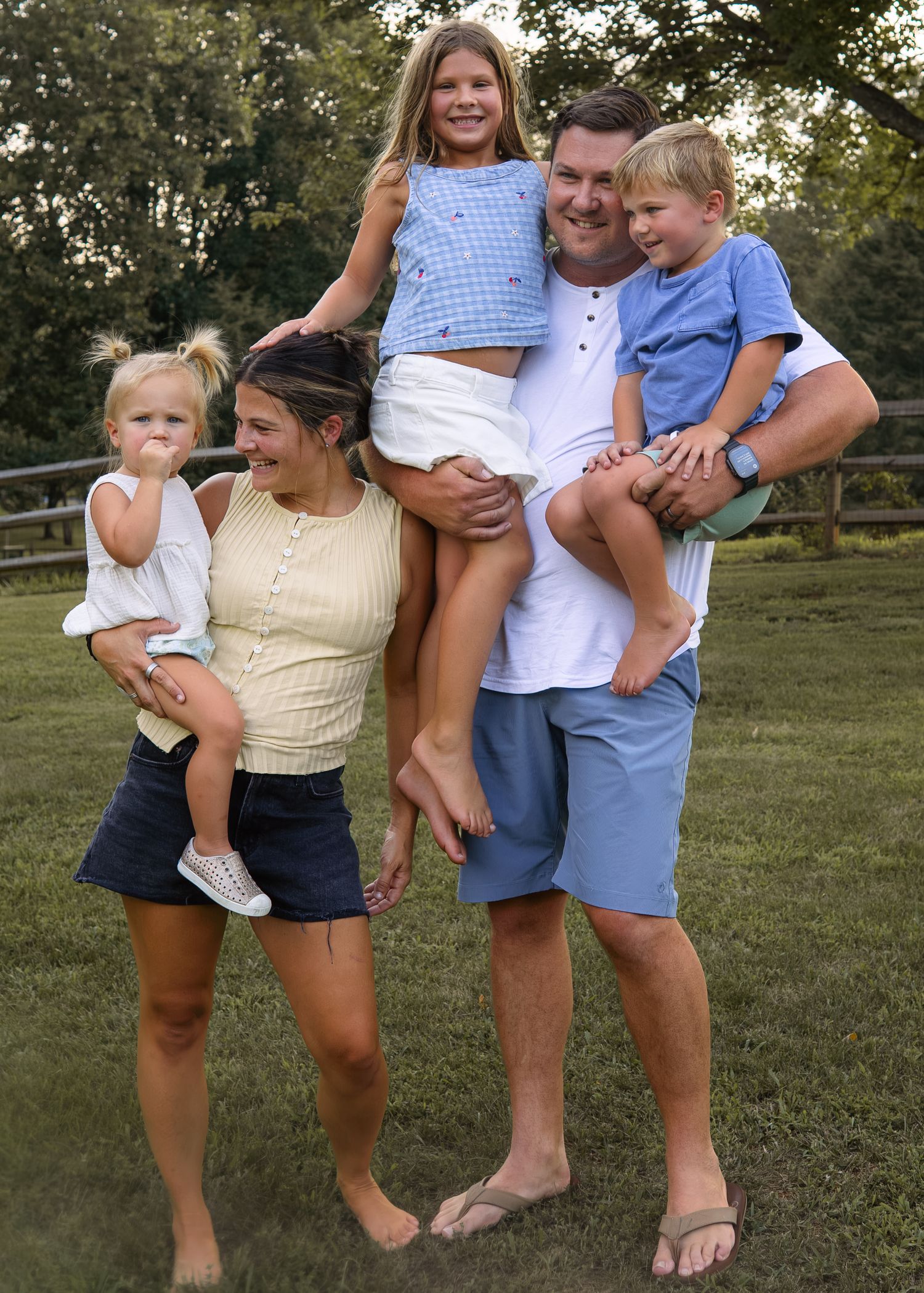 Happy family of five posing outdoors on green grass, parents holding three young children on a sunny day.