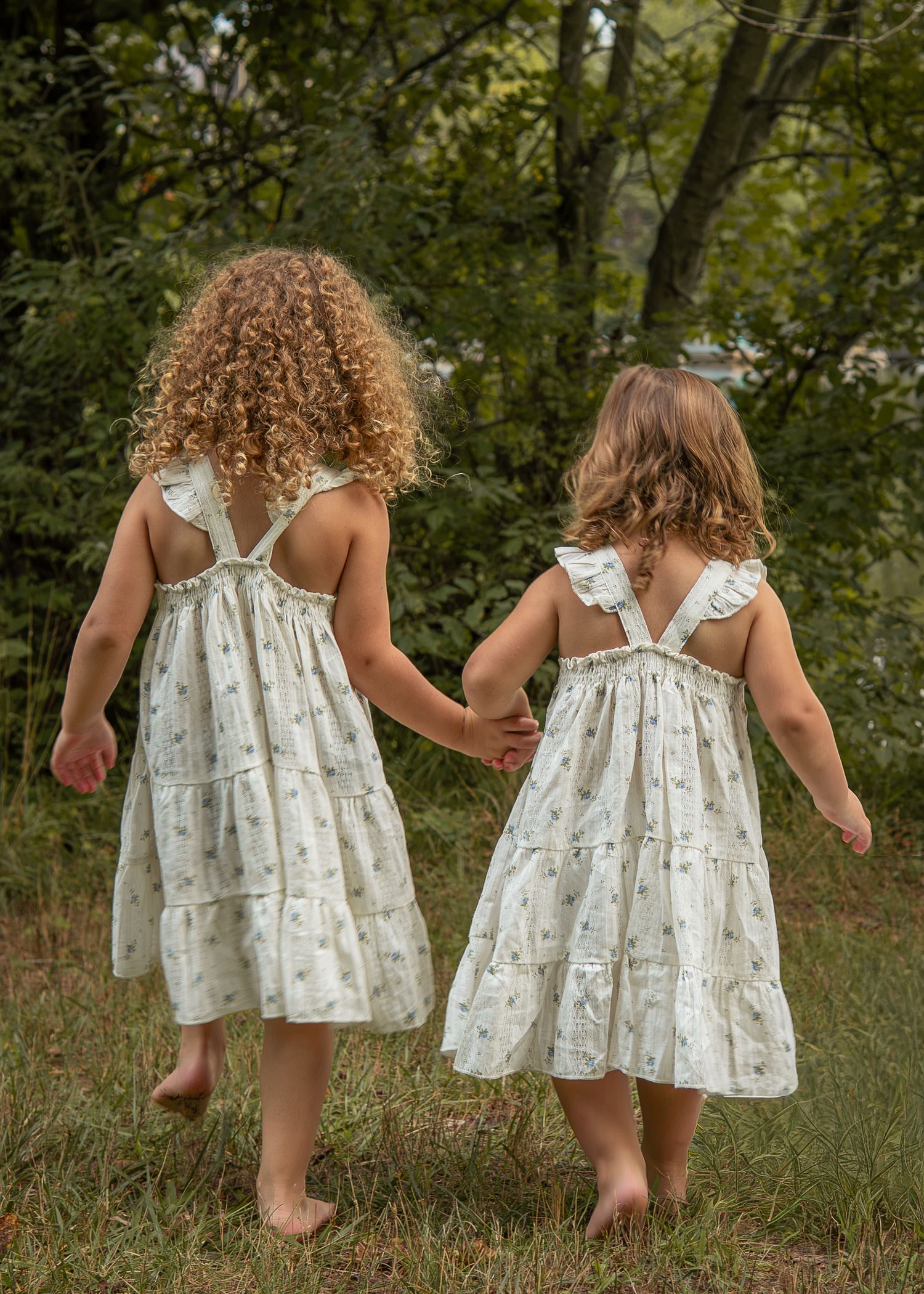Two young girls in matching white floral dresses holding hands while walking barefoot through green grass.