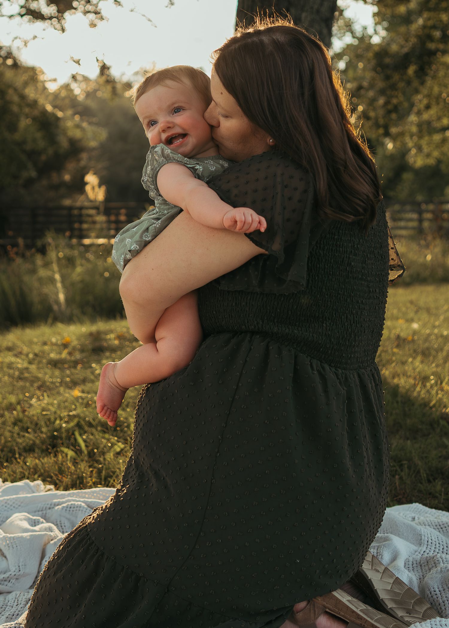 Mother in dark green dress kissing her smiling baby outdoors during golden hour on a blanket.