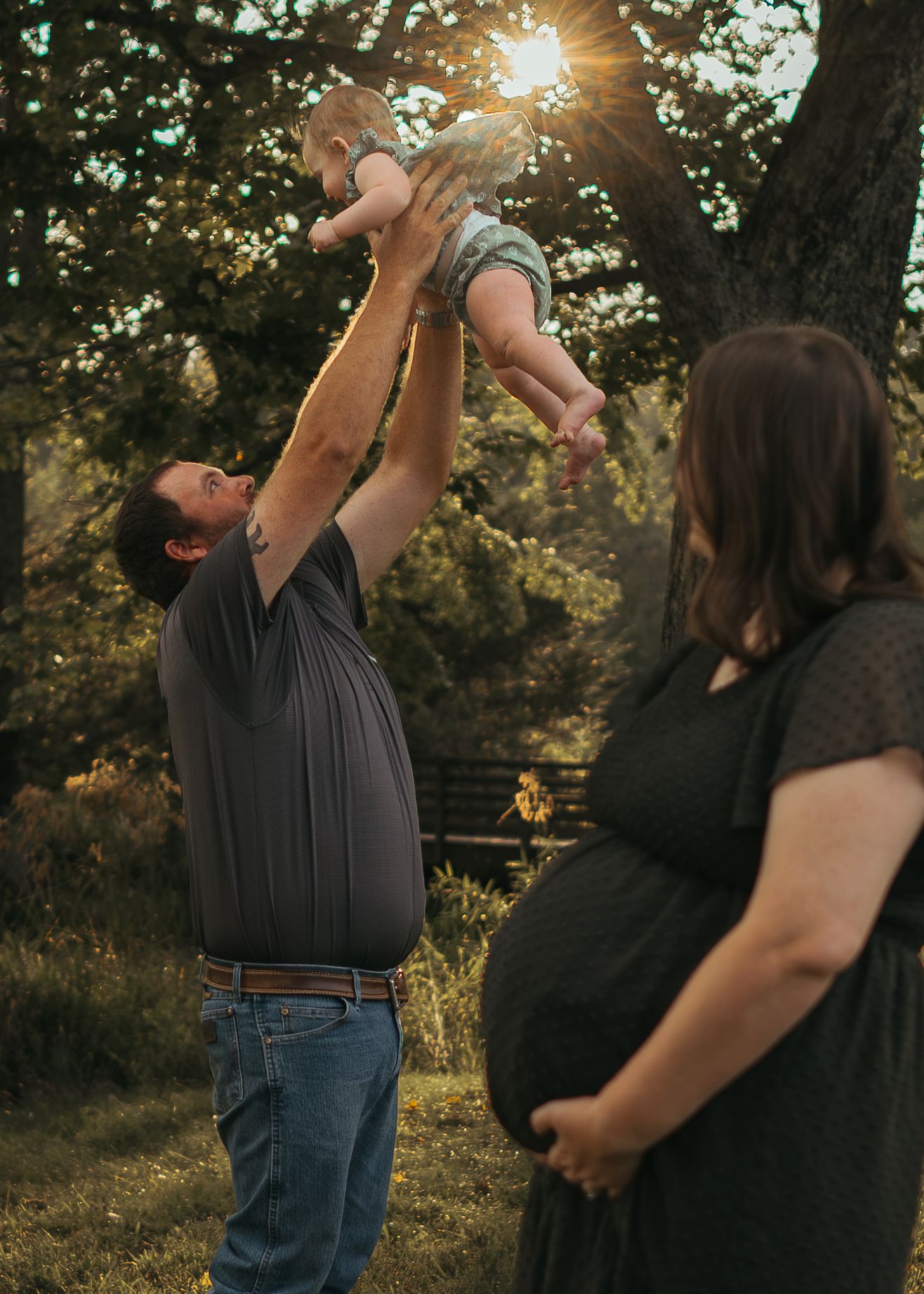A father lifts a baby in the air outdoors at golden hour while a pregnant woman stands nearby.