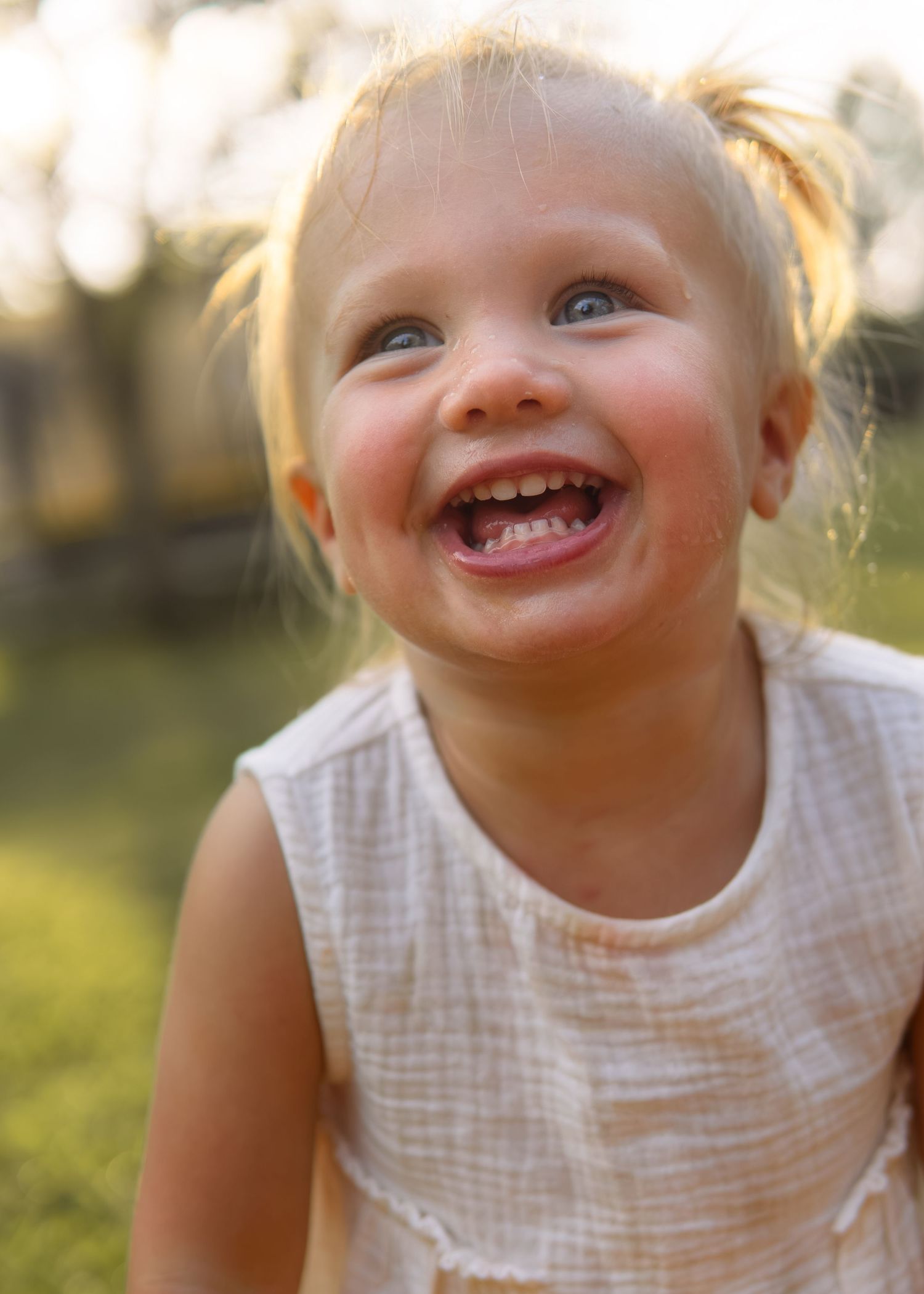 Smiling toddler girl with blonde pigtails and blue eyes wearing a white dress outdoors in golden sunlight.