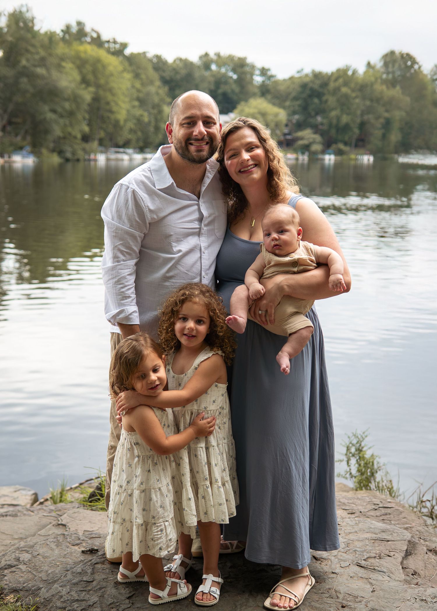 Family of five posing by a lake, parents with three young children, smiling outdoors near water with trees in background.