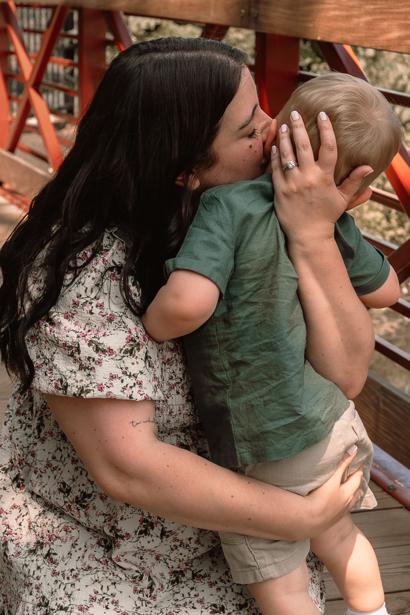 Loving mother with dark hair kissing young toddler boy on wooden bridge outdoors.