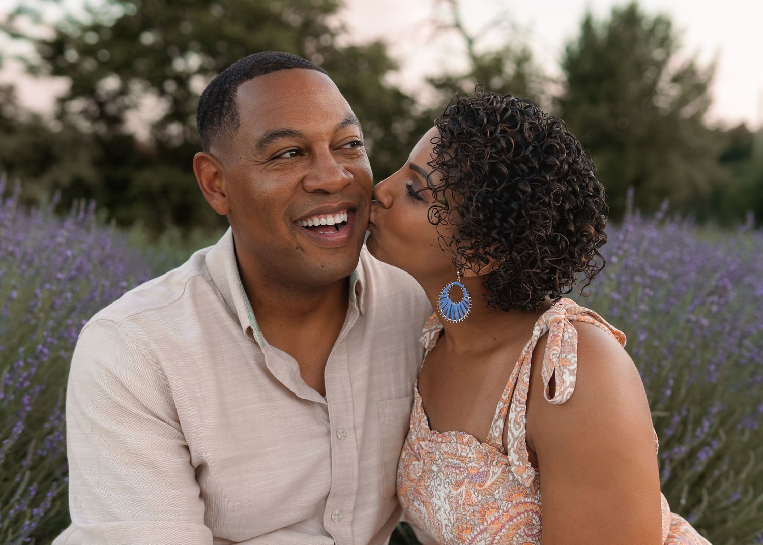Happy couple in lavender field, woman kissing mans cheek while he smiles broadly.