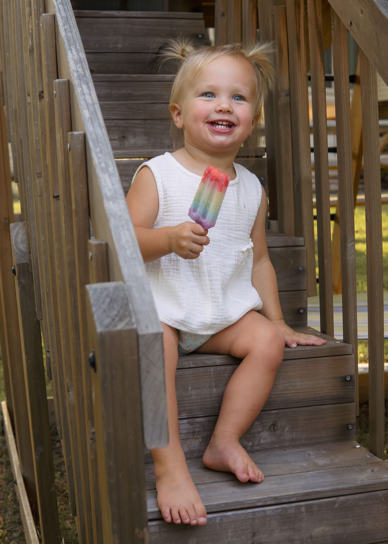 A happy blonde toddler girl sits on wooden steps eating a colorful rainbow popsicle outdoors.