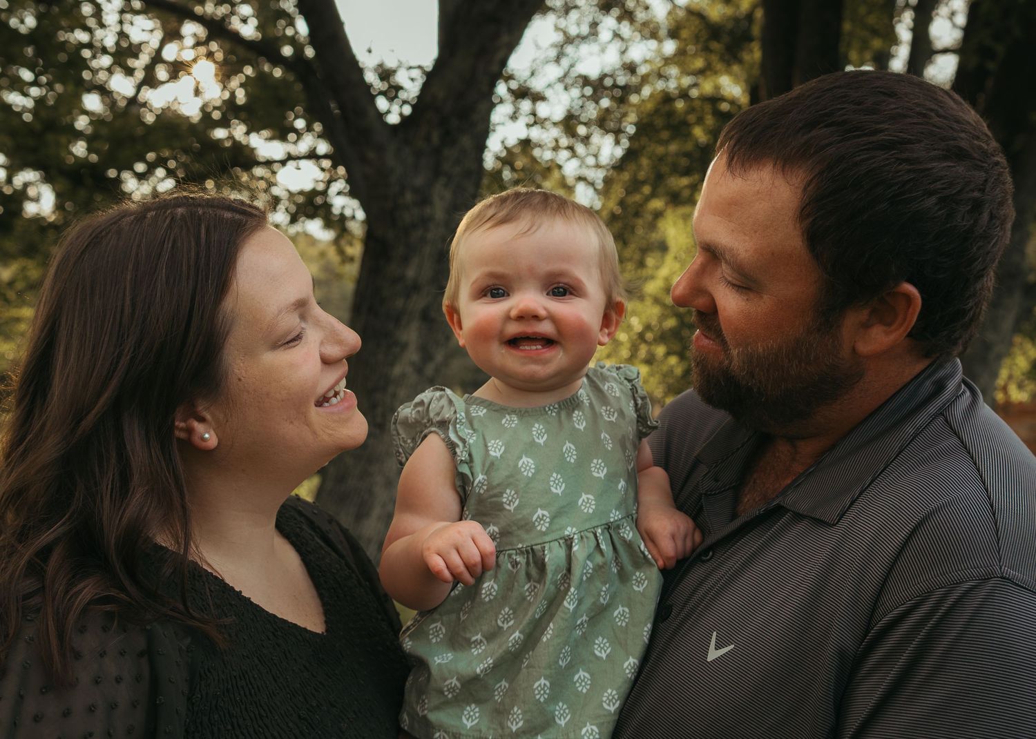 A young family smiling outdoors, parents holding their happy baby girl in a green dress among autumn trees.