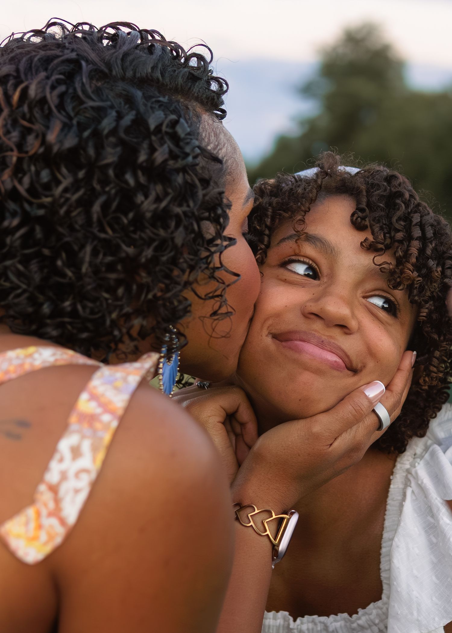 A woman lovingly kisses the cheek of a smiling young girl with curly hair outdoors.