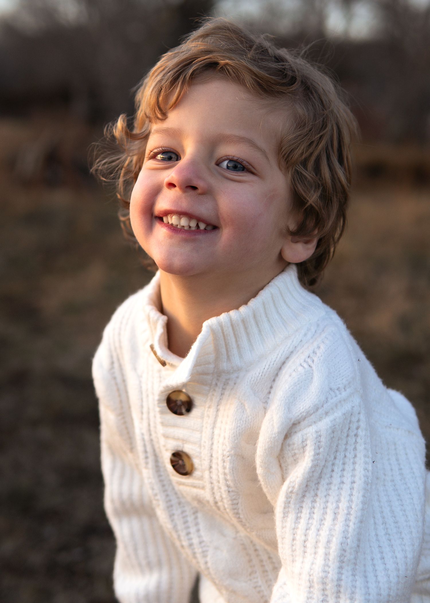 Happy toddler boy with curly blond hair smiling brightly, wearing a cream knit sweater outdoors.