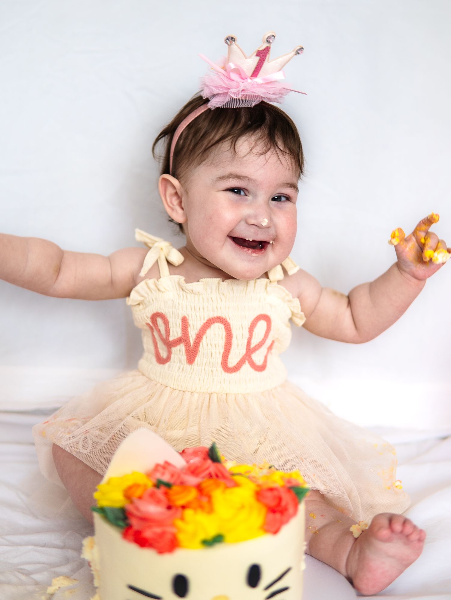 Baby in peach dress laughing near colorful birthday cake with flowers.