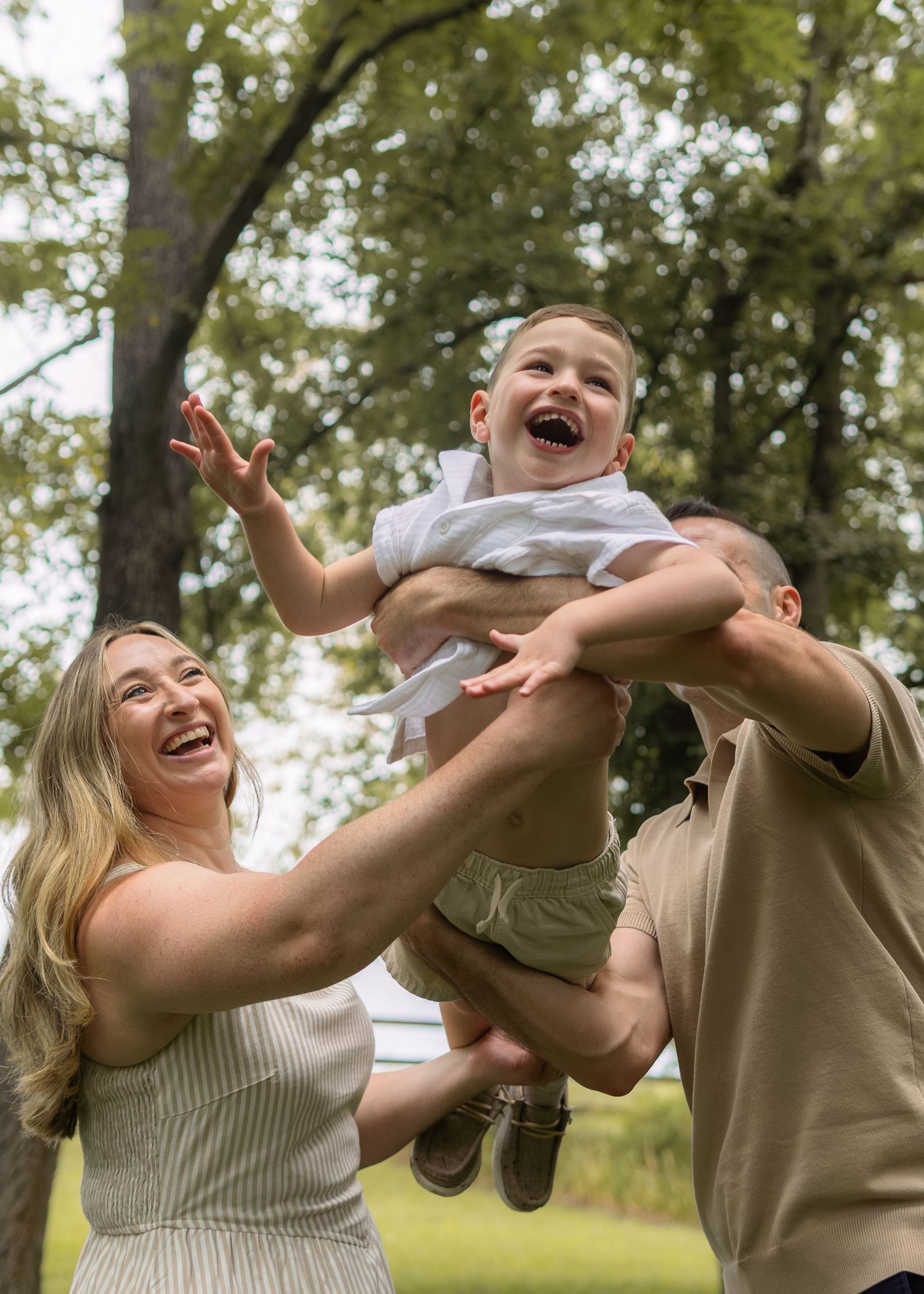 A joyful family laughs outdoors as parents lift their giggling toddler high in the air surrounded by lush green trees.