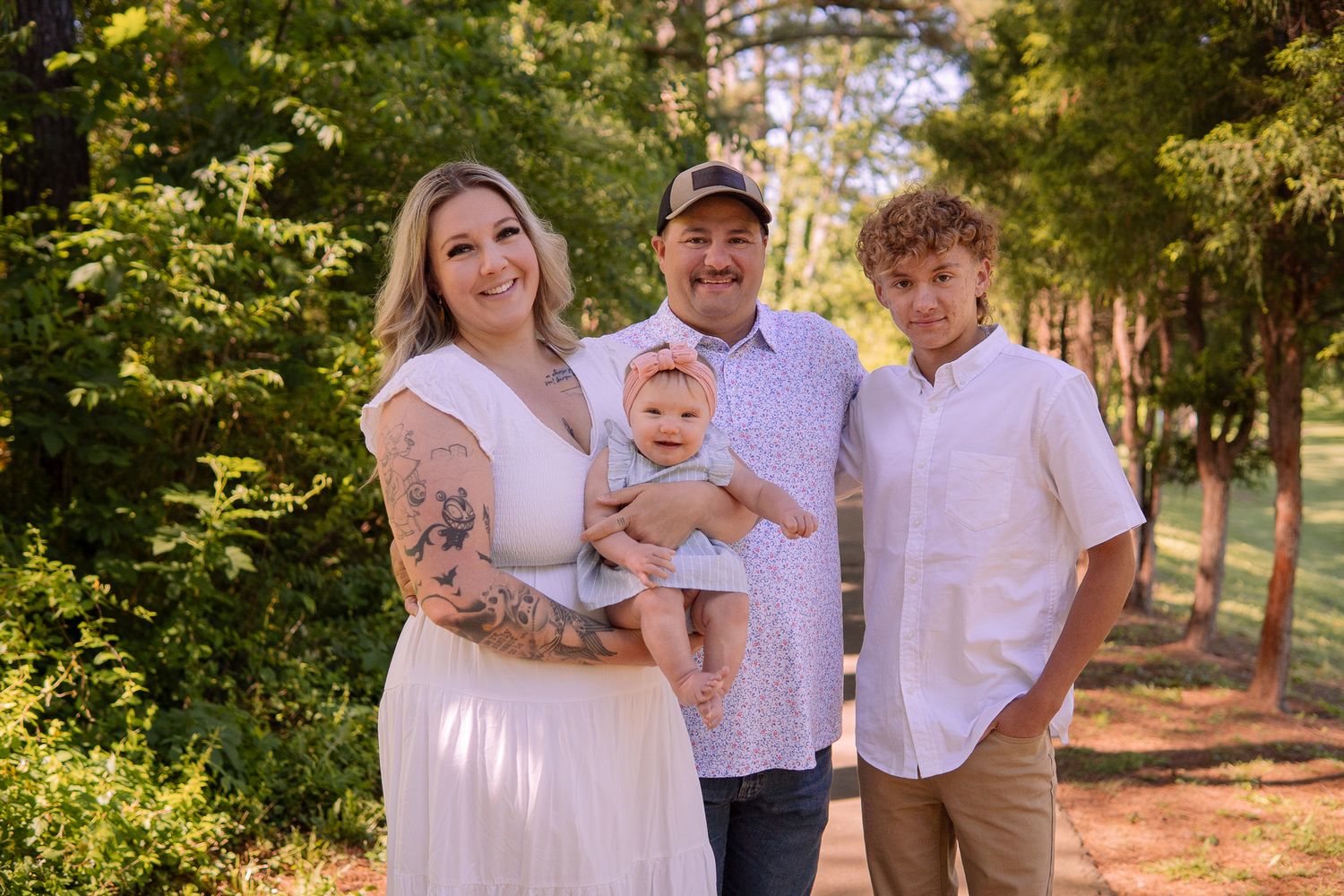 Outdoor family photo with coordinated white outfits against a backdrop of sunlit pine trees.