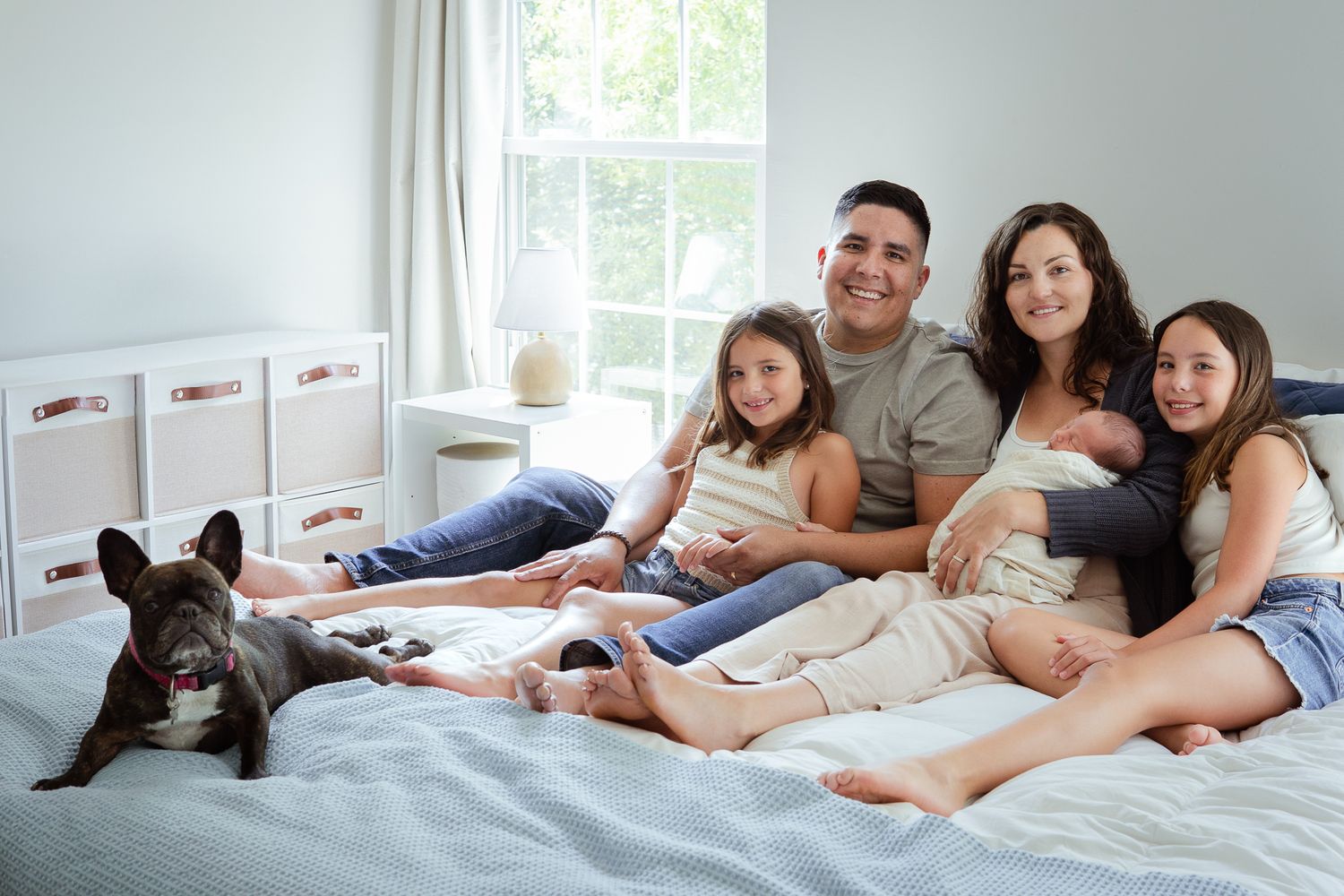Family relaxes on bed with newborn baby and French Bulldog pet.
