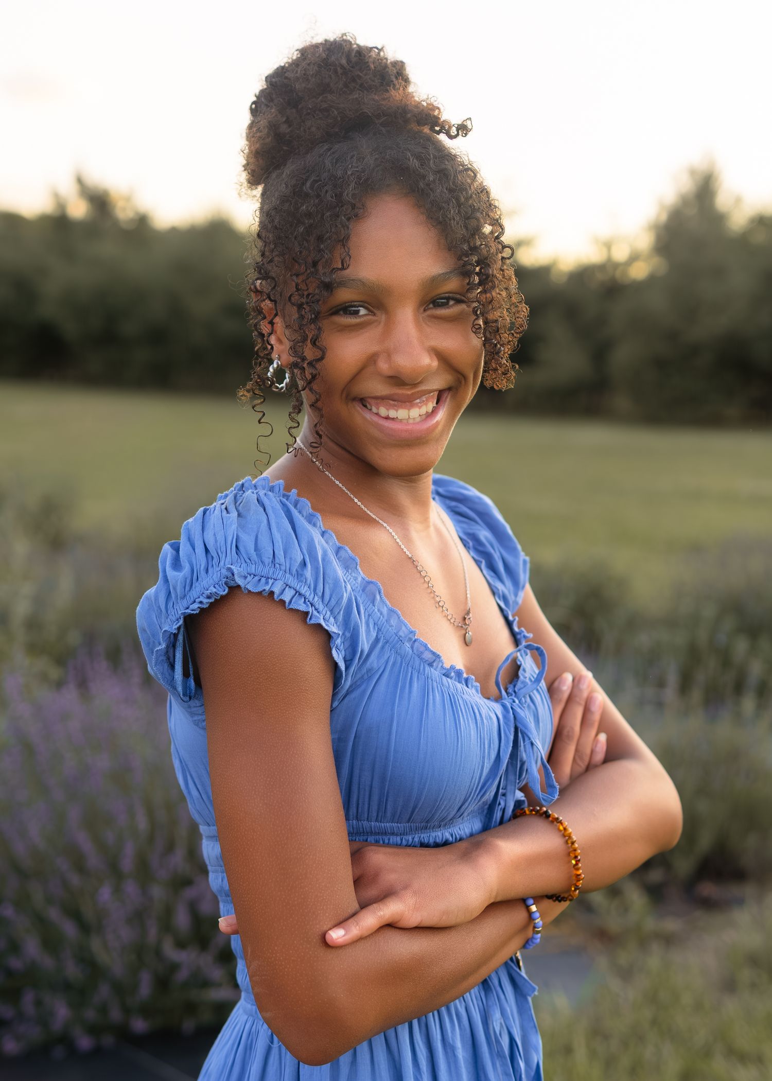 Teen Black girl in blue dress smiling with arms crossed in lavender field at golden hour.