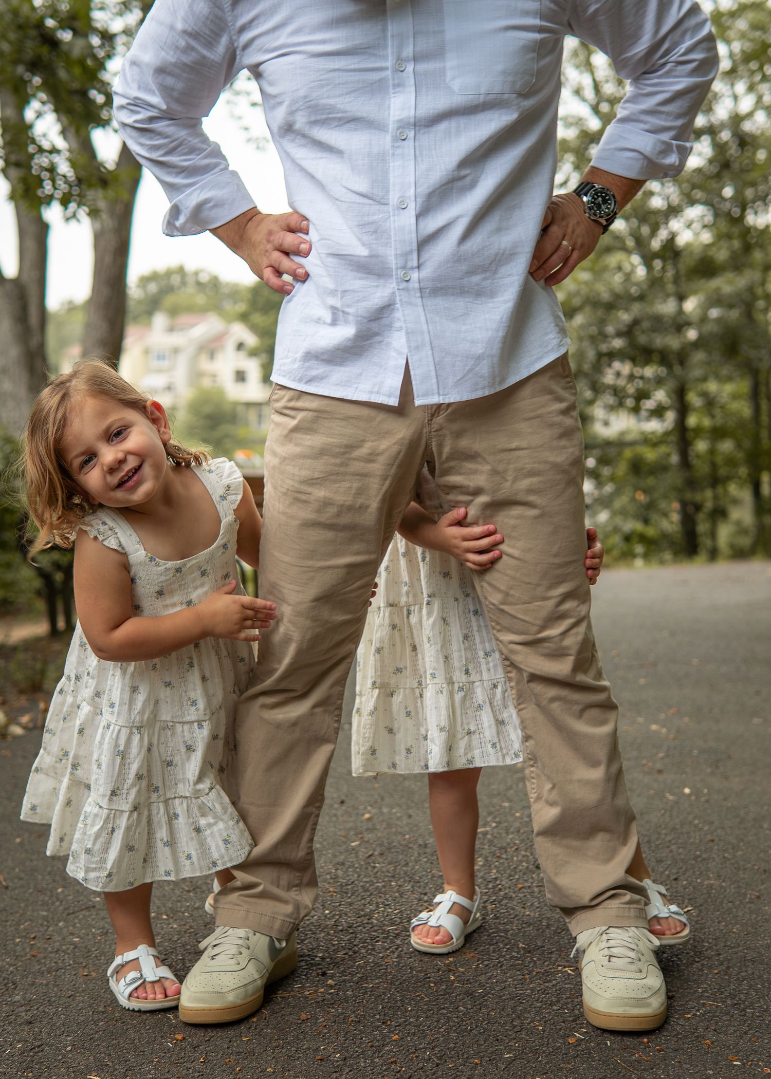 A young girl in a white floral dress smiles while standing between an adult's legs outdoors on a path.