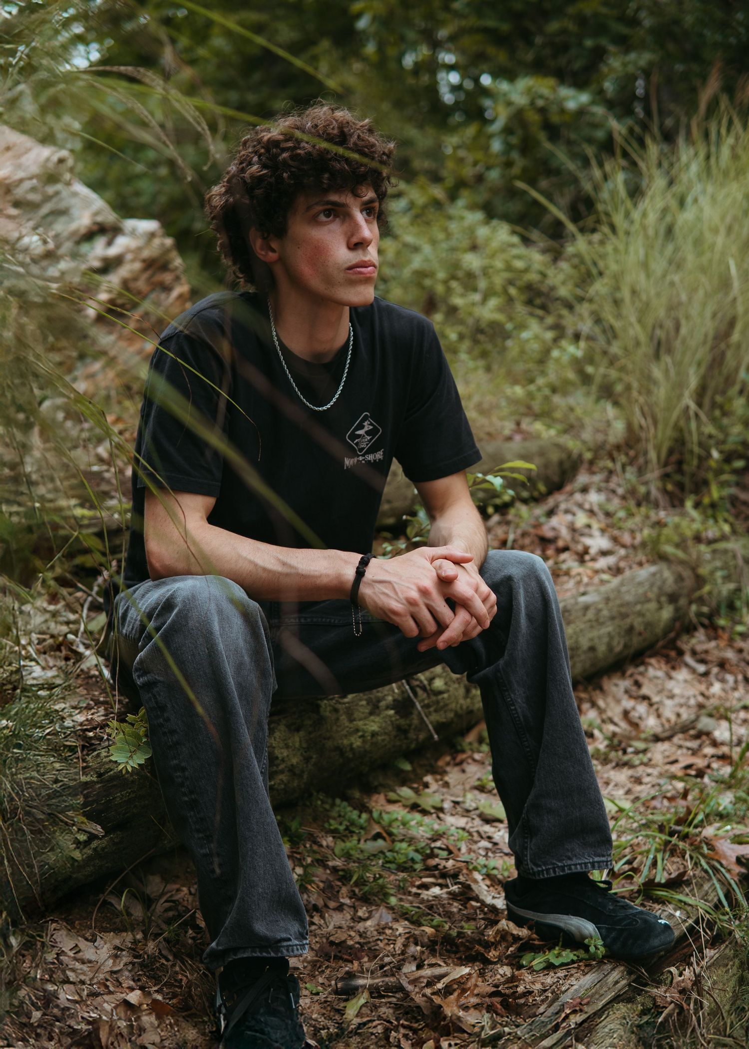 Young man with curly hair sits on a log in a forest, wearing a black t-shirt and gray jeans.