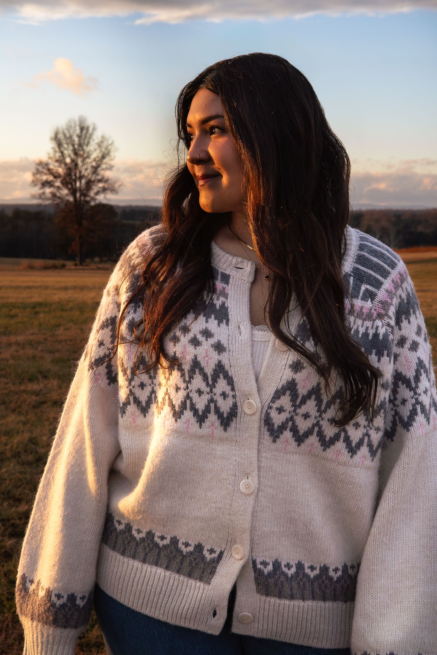 Young woman in Nordic patterned cardigan gazing into distance in open field at sunset.