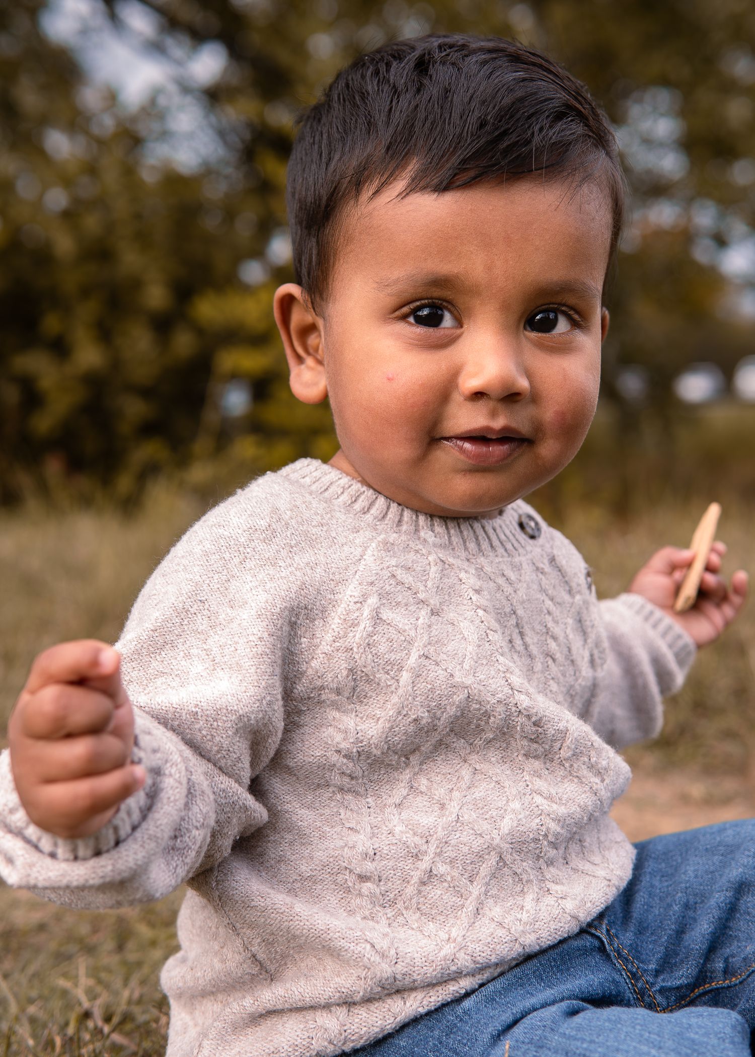 Cute baby boy in beige knit sweater sitting outdoors holding a small stick.