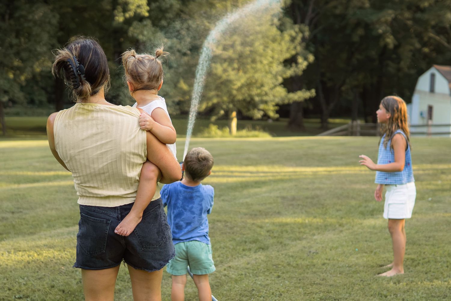 A mother holds her toddler while two children watch a garden hose sprinkler in a sunny yard.