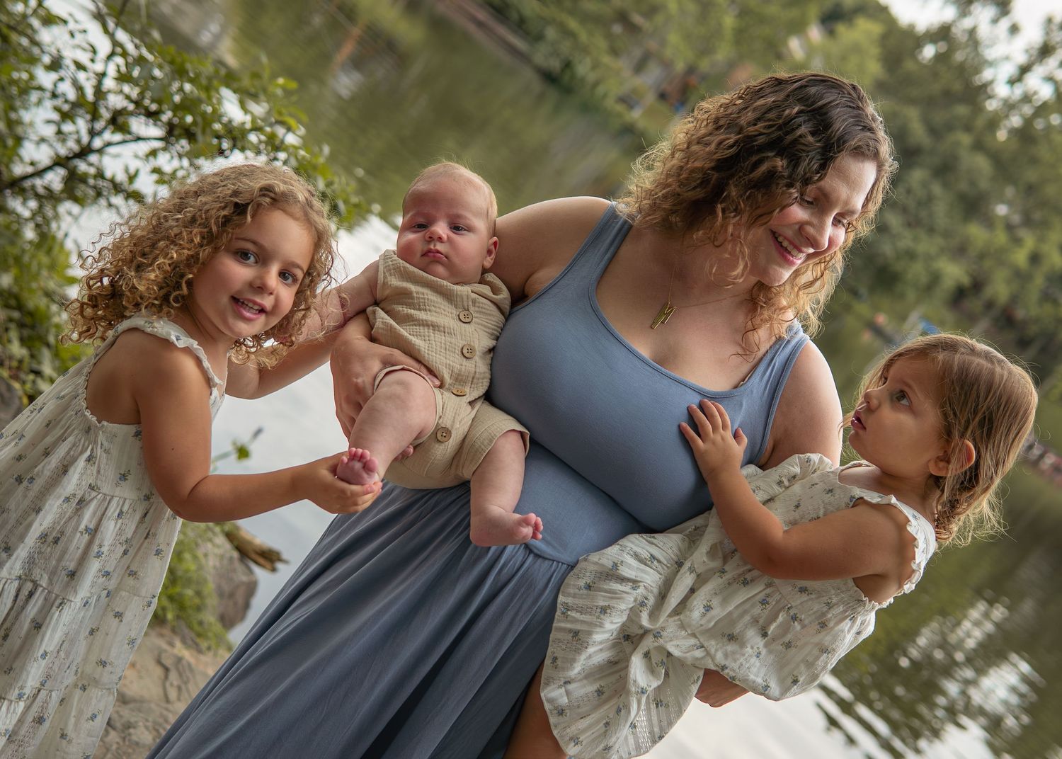 A mother smiles holding her baby while two young curly-haired girls stand beside her outdoors near a lake.