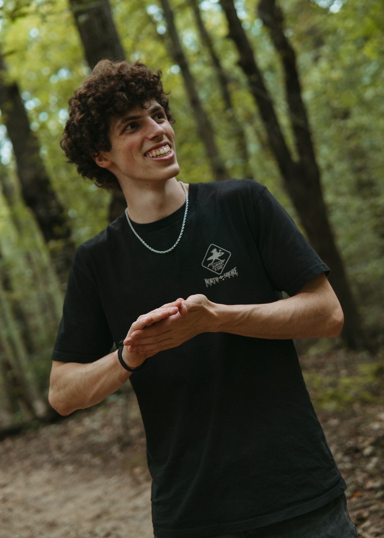 Smiling young man with curly hair wearing a black graphic t-shirt and silver chain necklace in a forest setting.