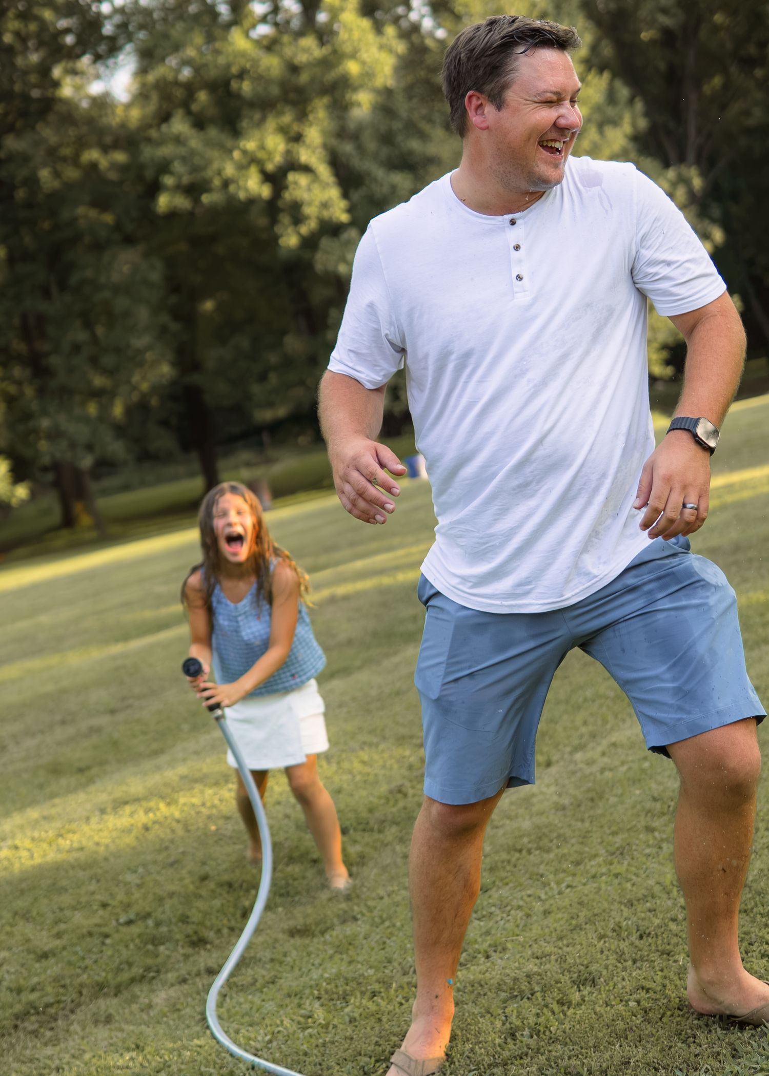 A laughing father runs as his daughter playfully sprays him with a garden hose in a green backyard.
