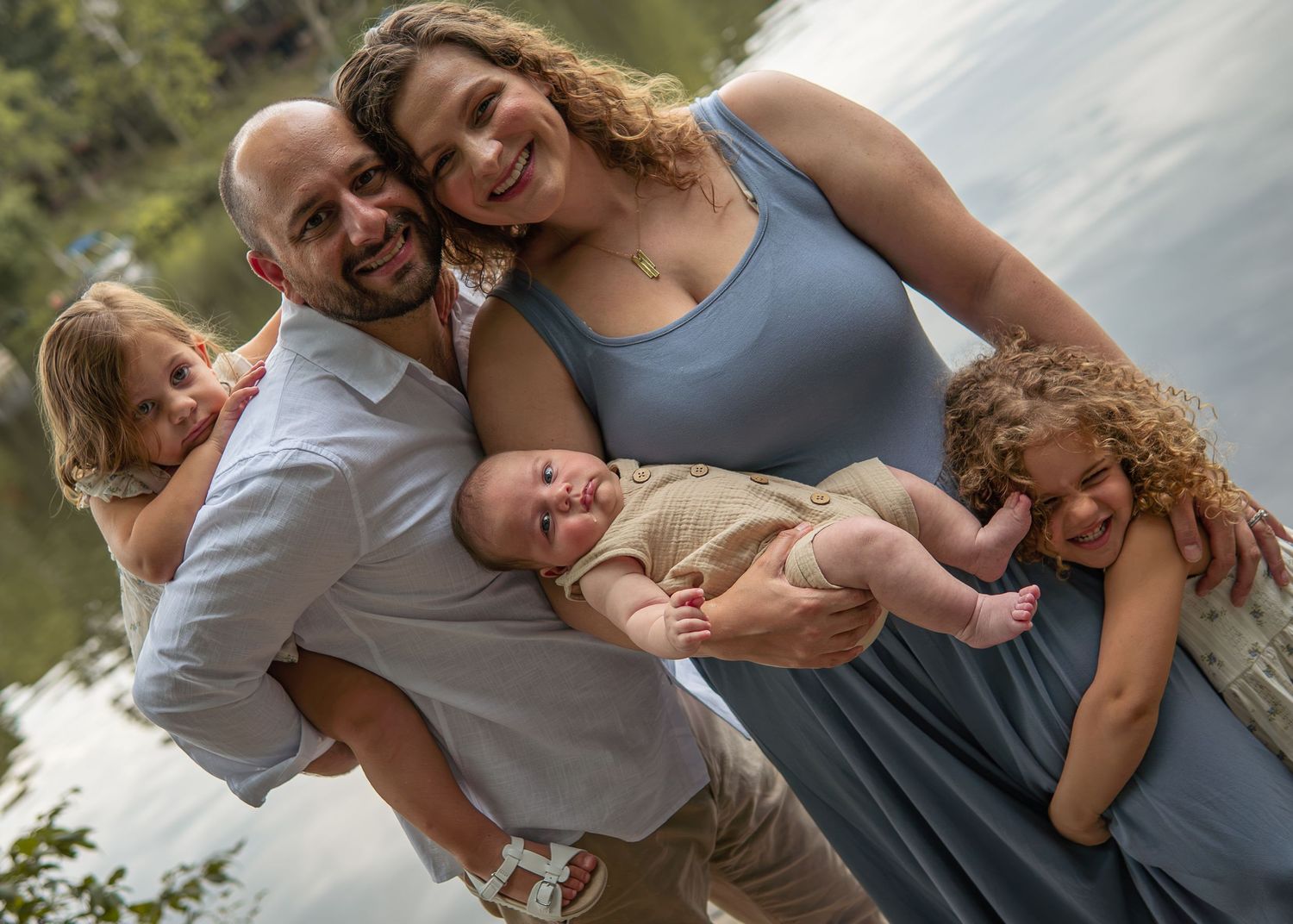 Happy family of five posing outdoors near a lake, parents holding three young children including a baby.