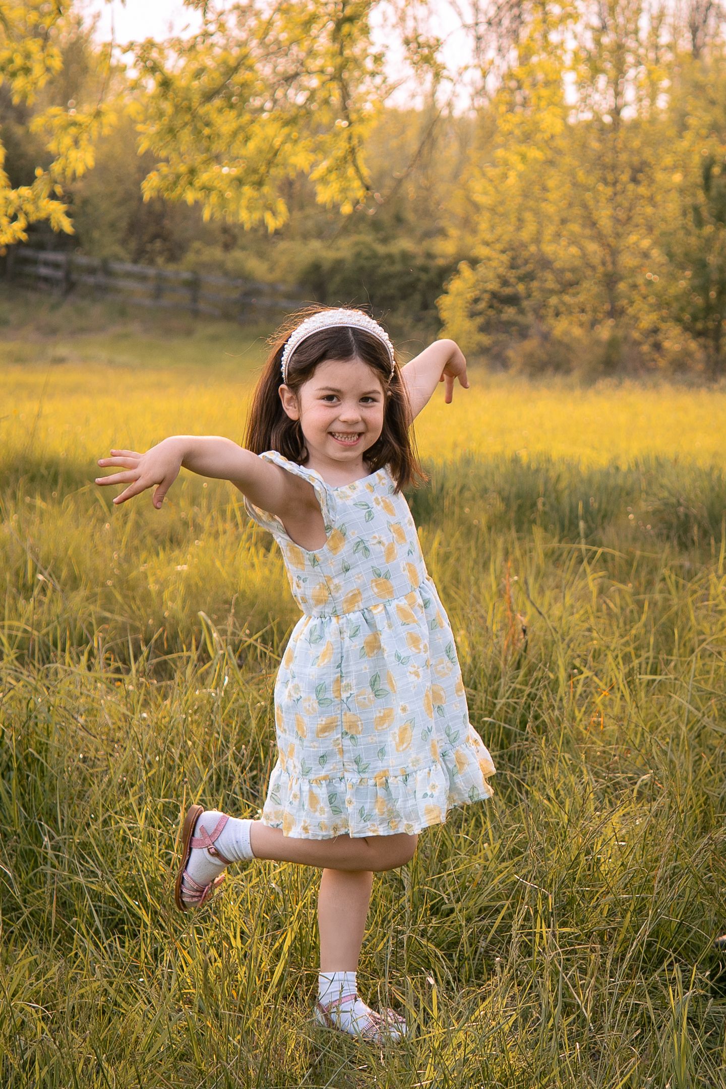 A child in a white floral dress dances playfully in a golden meadow at sunset with arms outstretched.