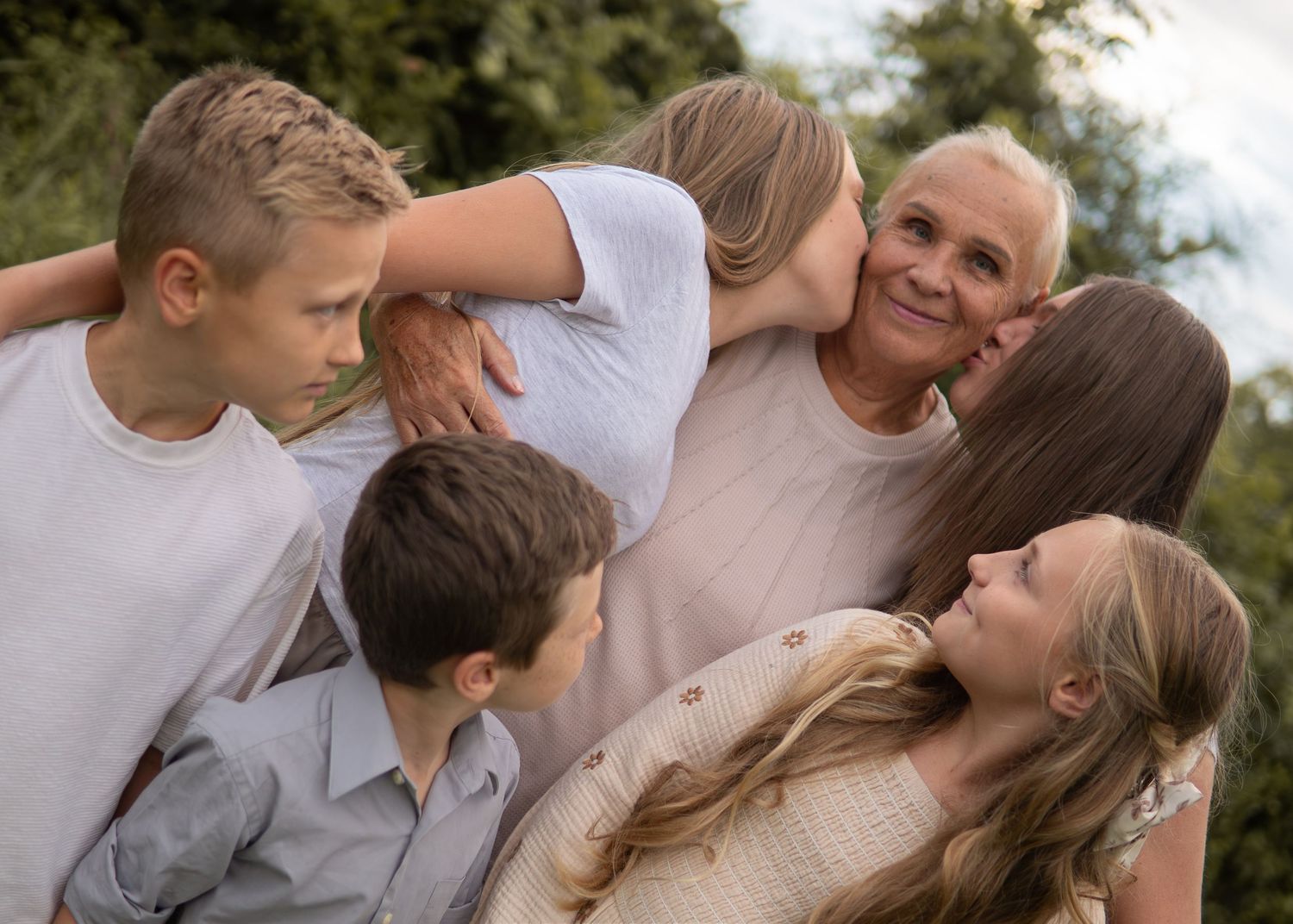 Multi-generational family group hug outdoors, grandparent surrounded by children and grandchildren smiling together.