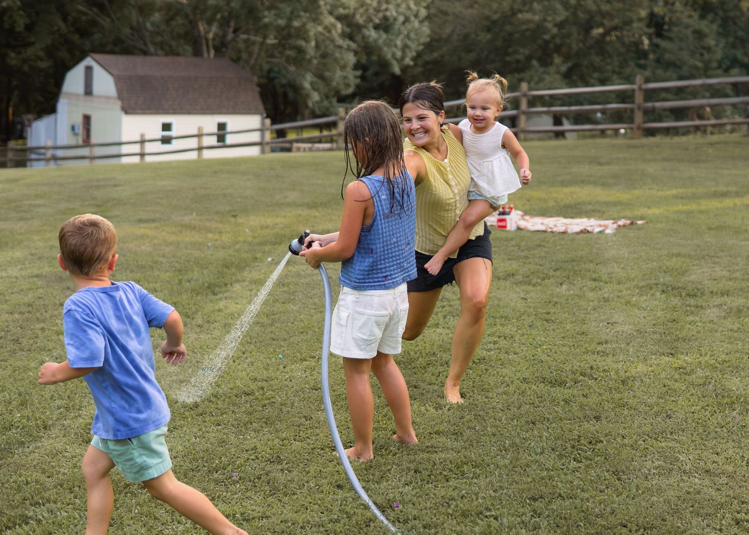 A woman and children laugh while playing with a water hose on a grassy backyard lawn.