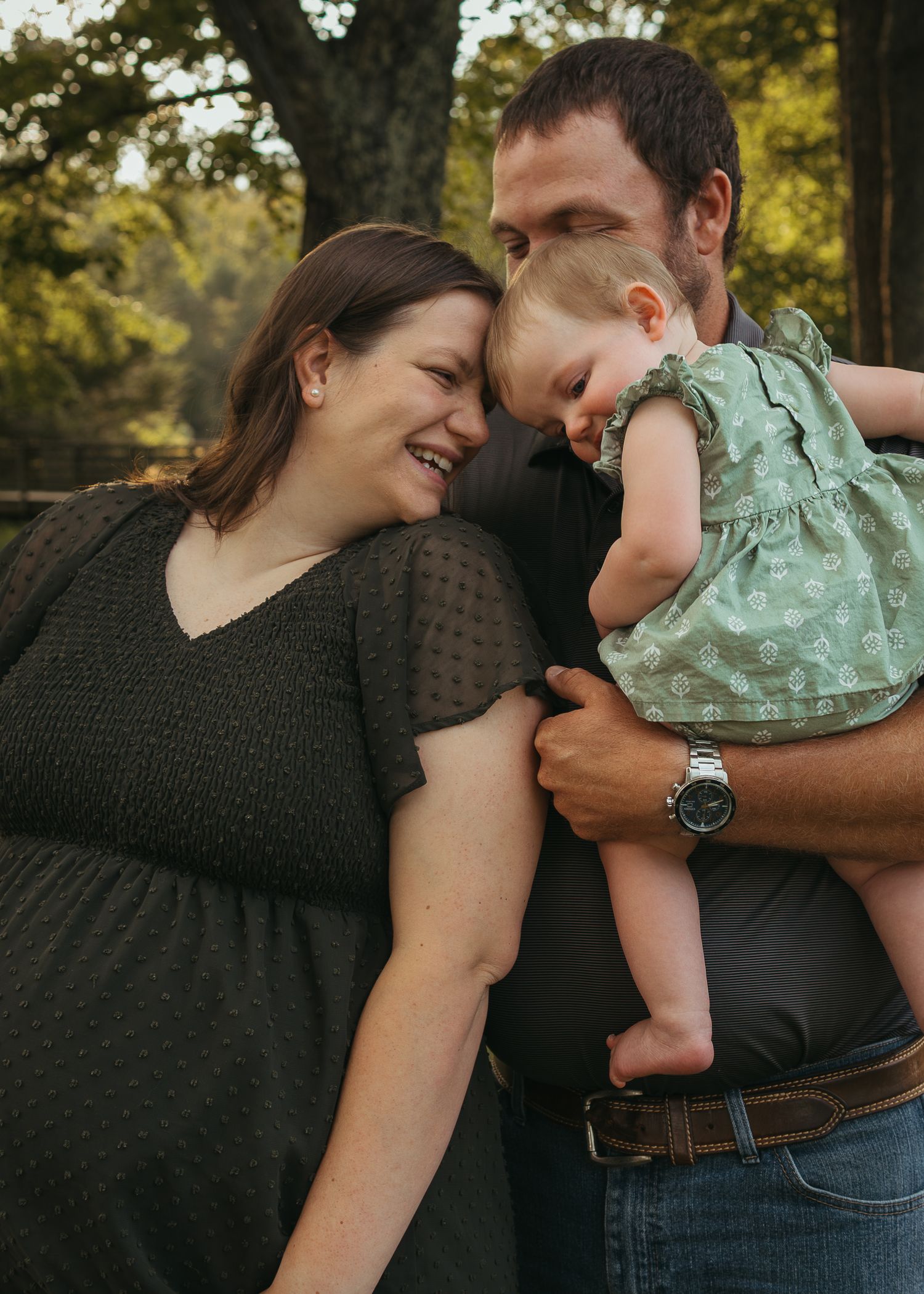 Pregnant woman and man holding baby girl outdoors, sharing a joyful family moment under trees.