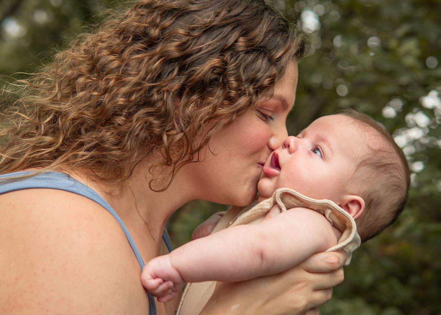 A curly-haired mother lovingly kisses her newborn baby outdoors with green trees in the background.