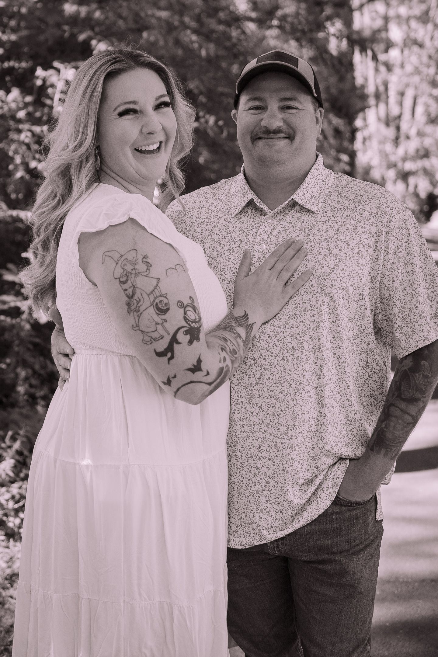 A black and white portrait of a couple outdoors showing off her arm tattoo in casual attire.