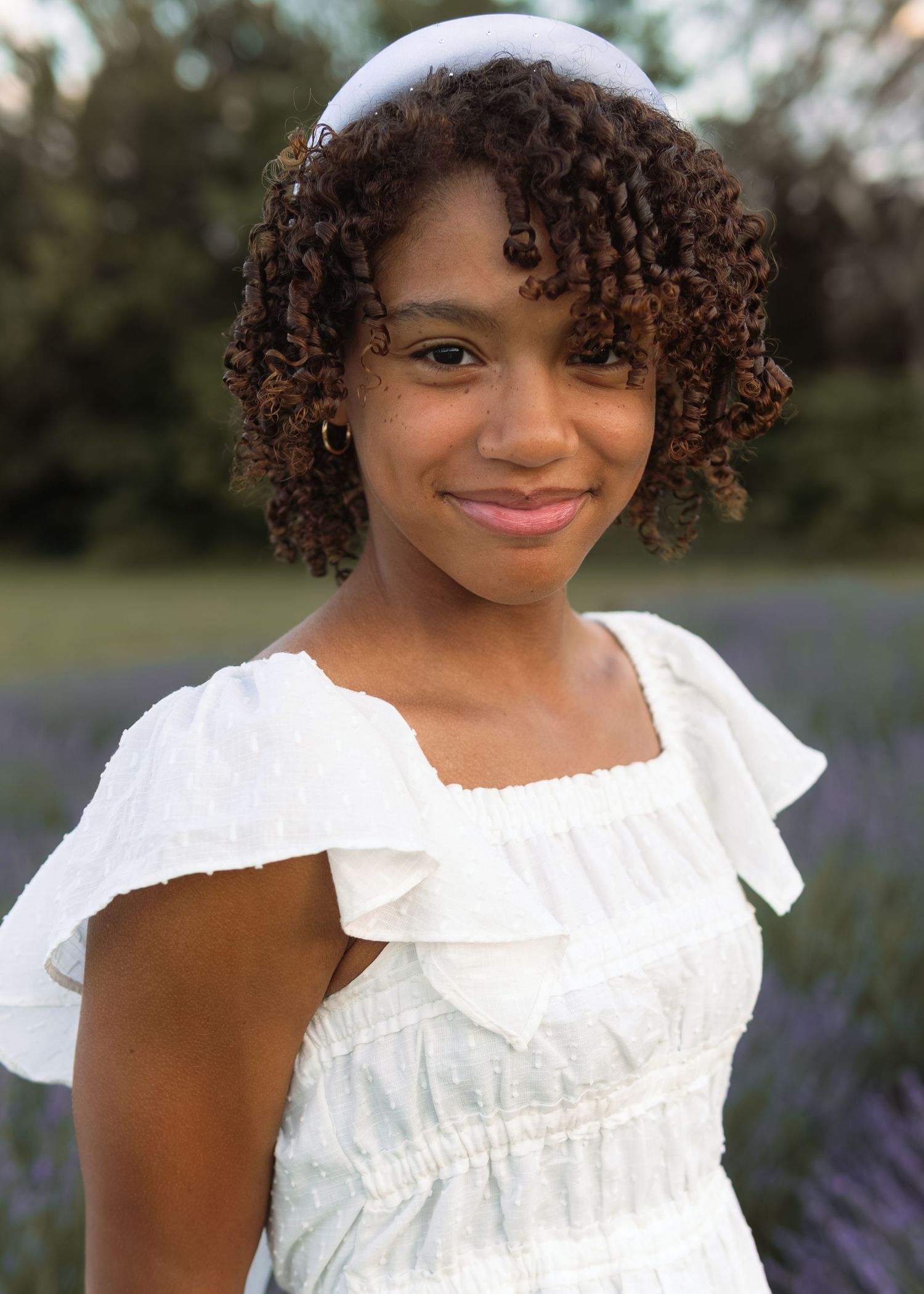 Young Black teen in white ruffled dress with headband smiling softly in a lavender field.