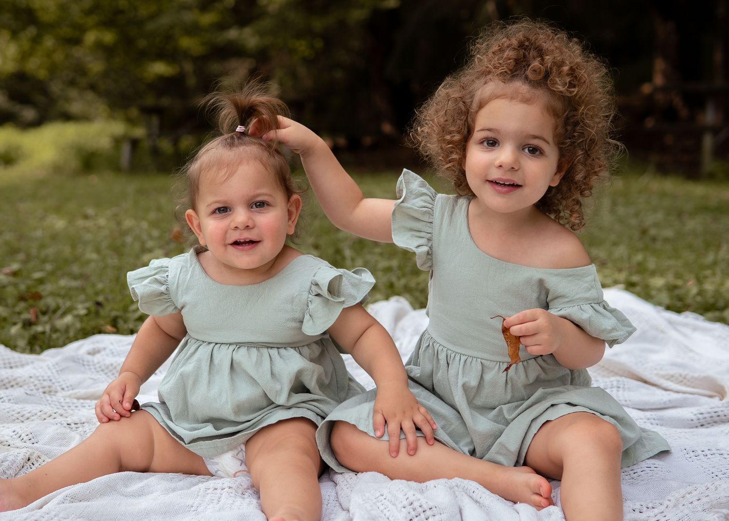 Two toddler sisters in matching sage green outfits sitting on blanket outdoors.