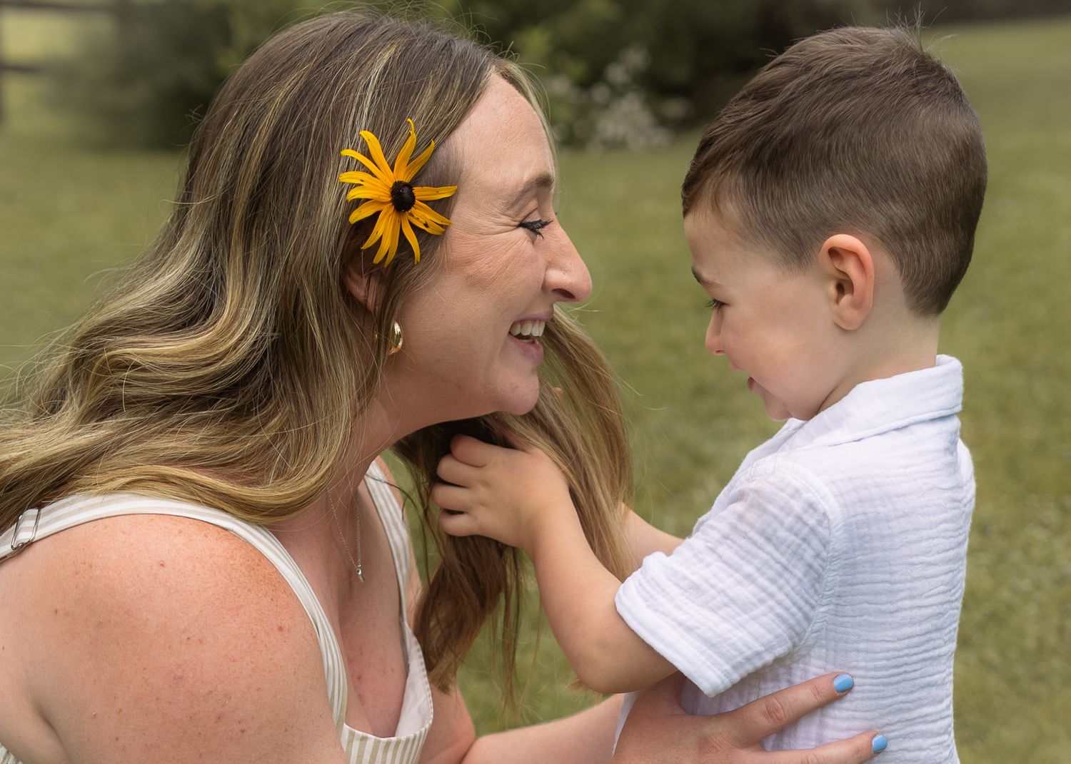 A smiling woman with a yellow flower in her hair lovingly holds a young boy outdoors in a green field.