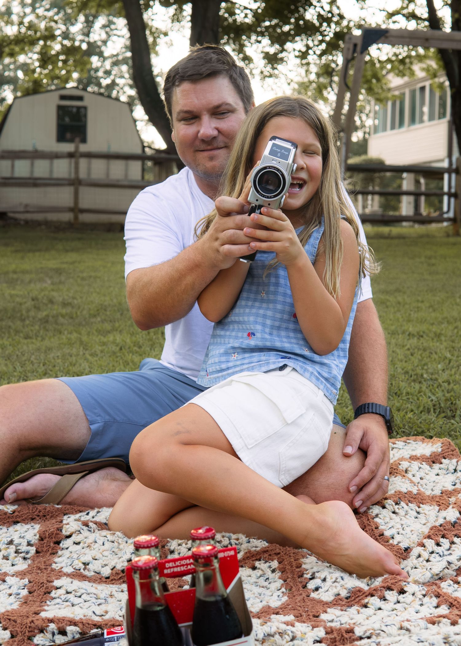 Man and girl sitting on picnic blanket outdoors, holding vintage camera with Coca-Cola bottles in foreground.