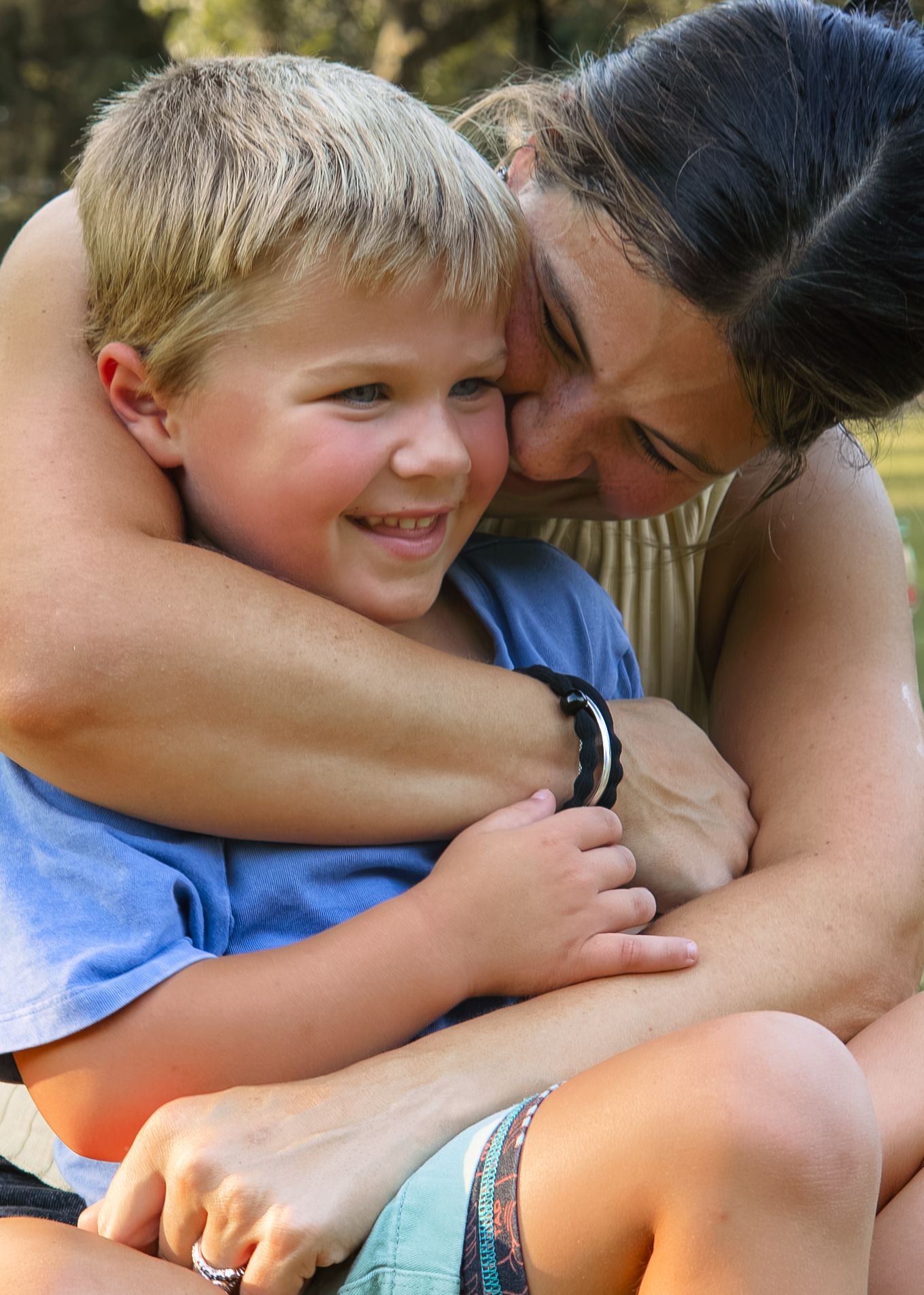 A woman hugs a smiling young boy outdoors, sharing a warm and loving embrace in a sunny park setting.