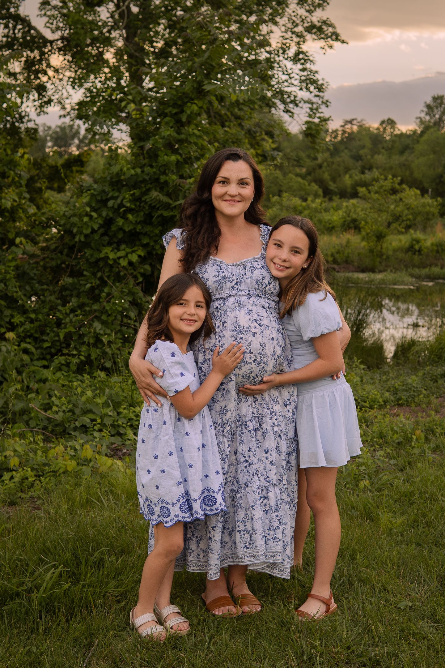 A family poses together outdoors at sunset, wearing coordinated blue and white dresses against a natural green backdrop.