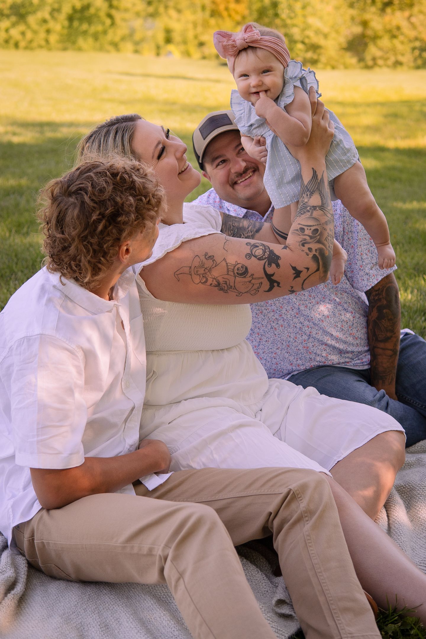 A joyful family moment captured on a blanket outdoors during golden hour with a baby in white.