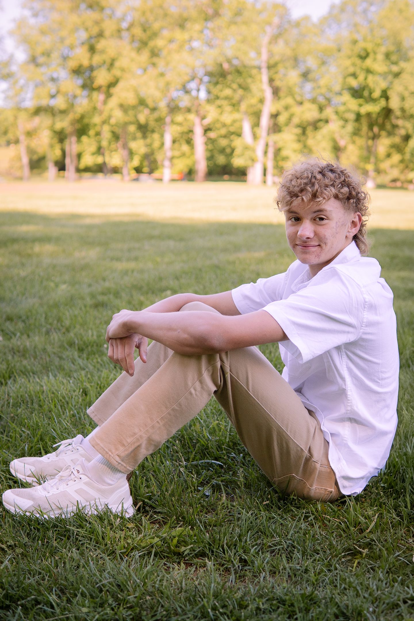 A casual portrait of a young person sitting on grass wearing a white shirt and khaki pants.