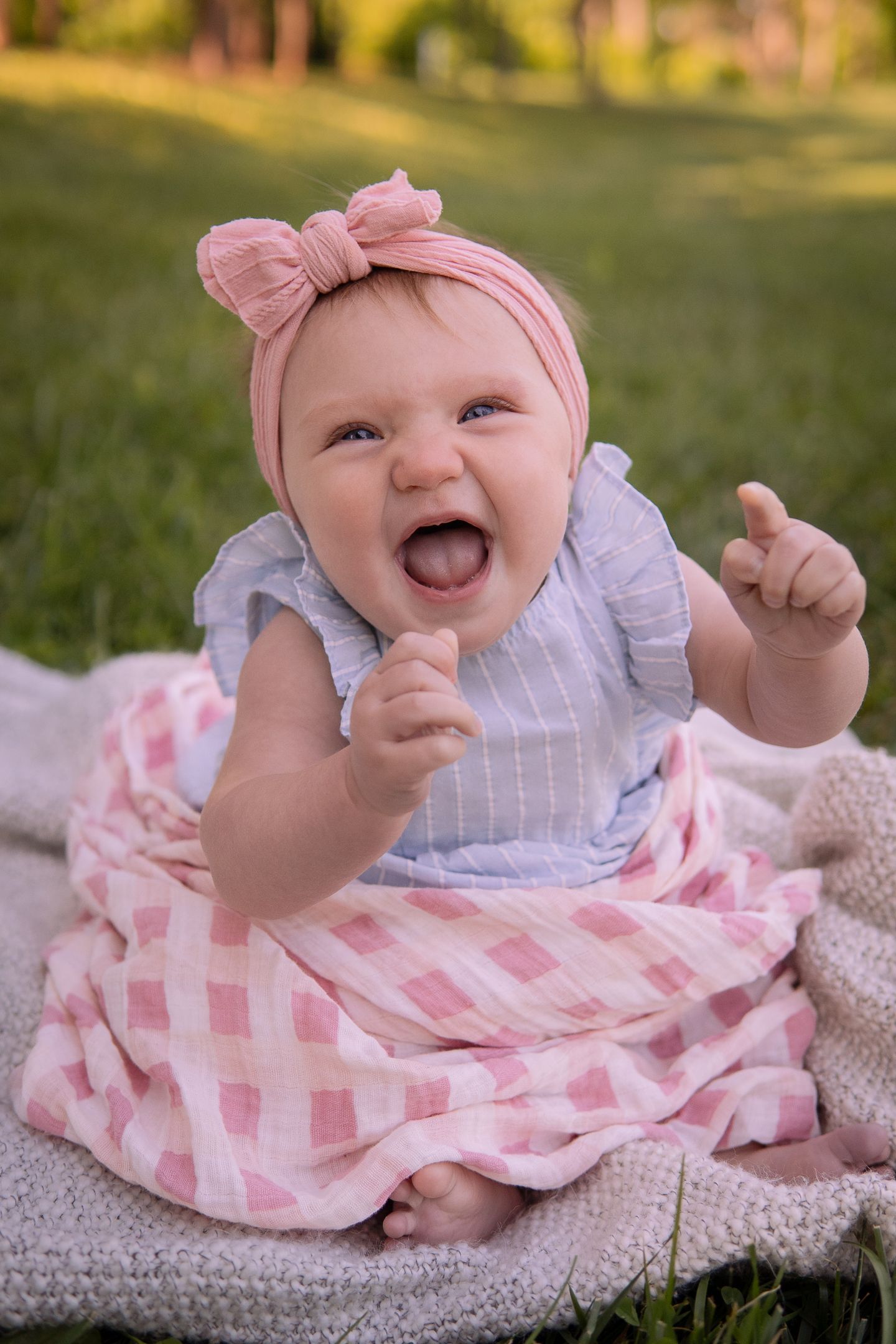 A happy baby wearing a pink bow headband and gingham dress smiles while sitting on a blanket outdoors.