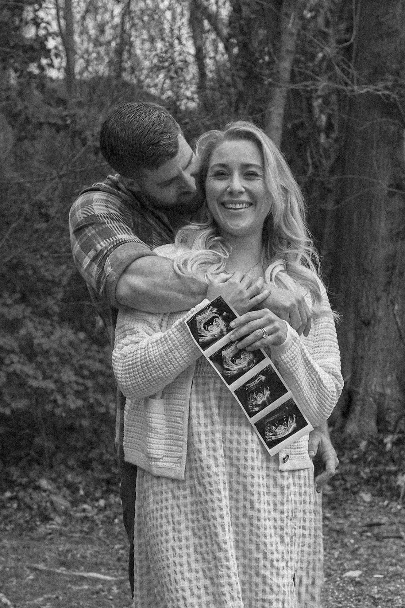 A couple embraces in a forest setting while holding ultrasound photos, announcing their pregnancy in a black and white photo.
