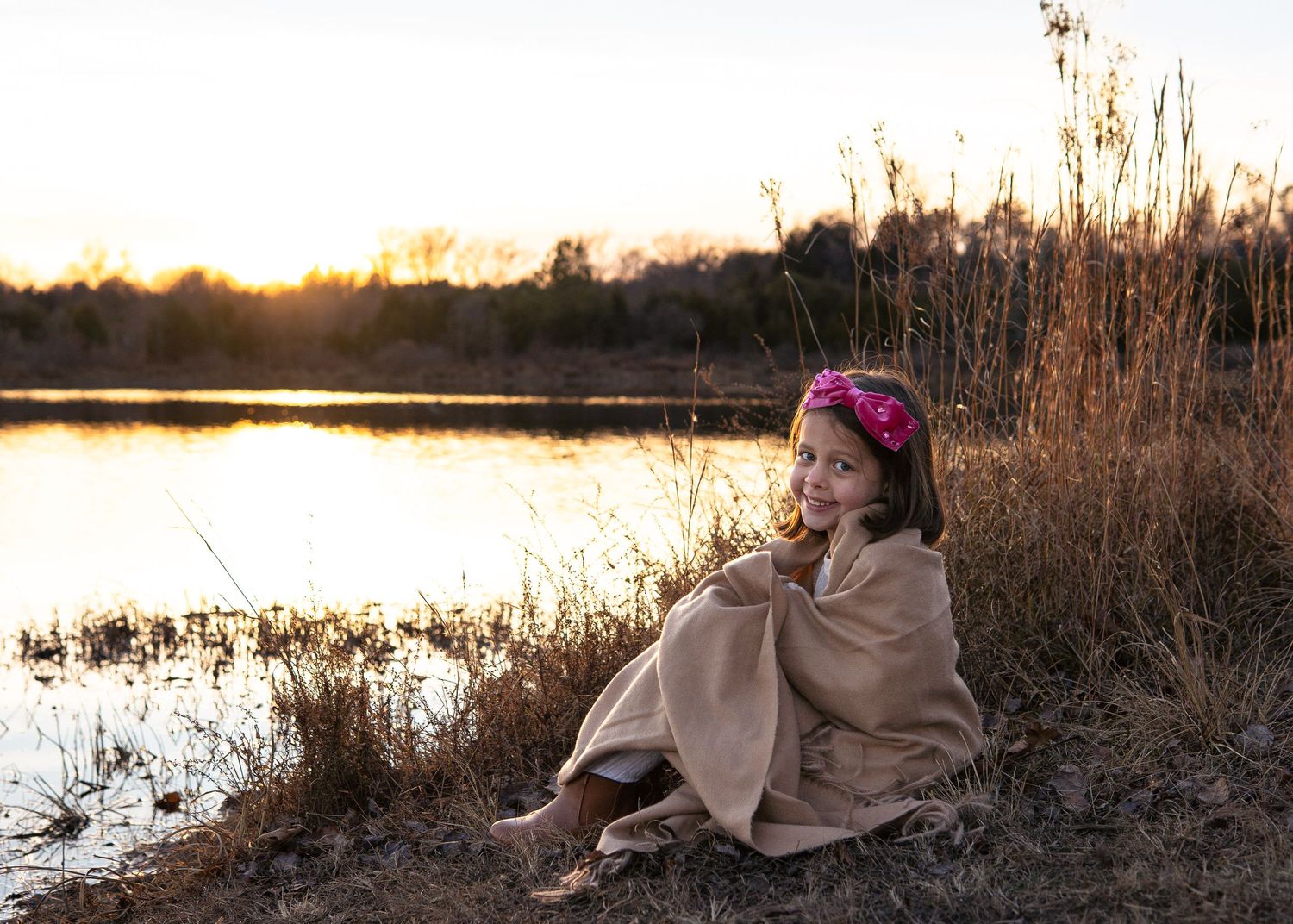 Young girl wrapped in a tan blanket with a pink floral headband, sitting by a lake at golden sunset.