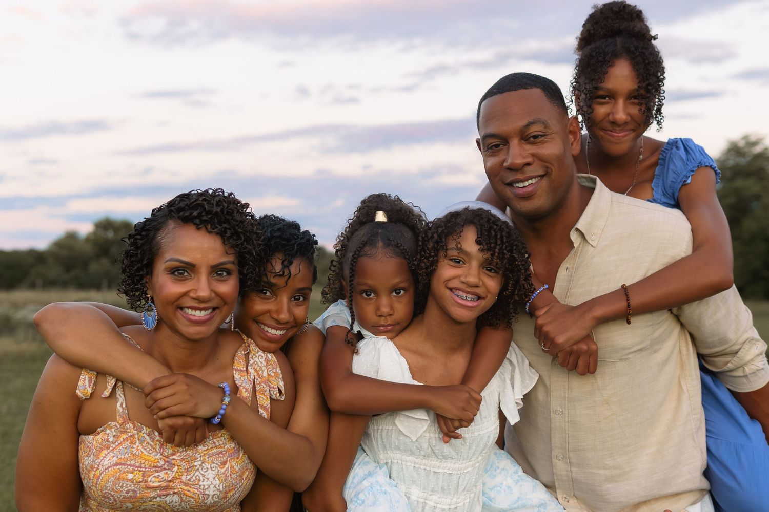 A joyful family of six poses together outdoors under a colorful evening sky.