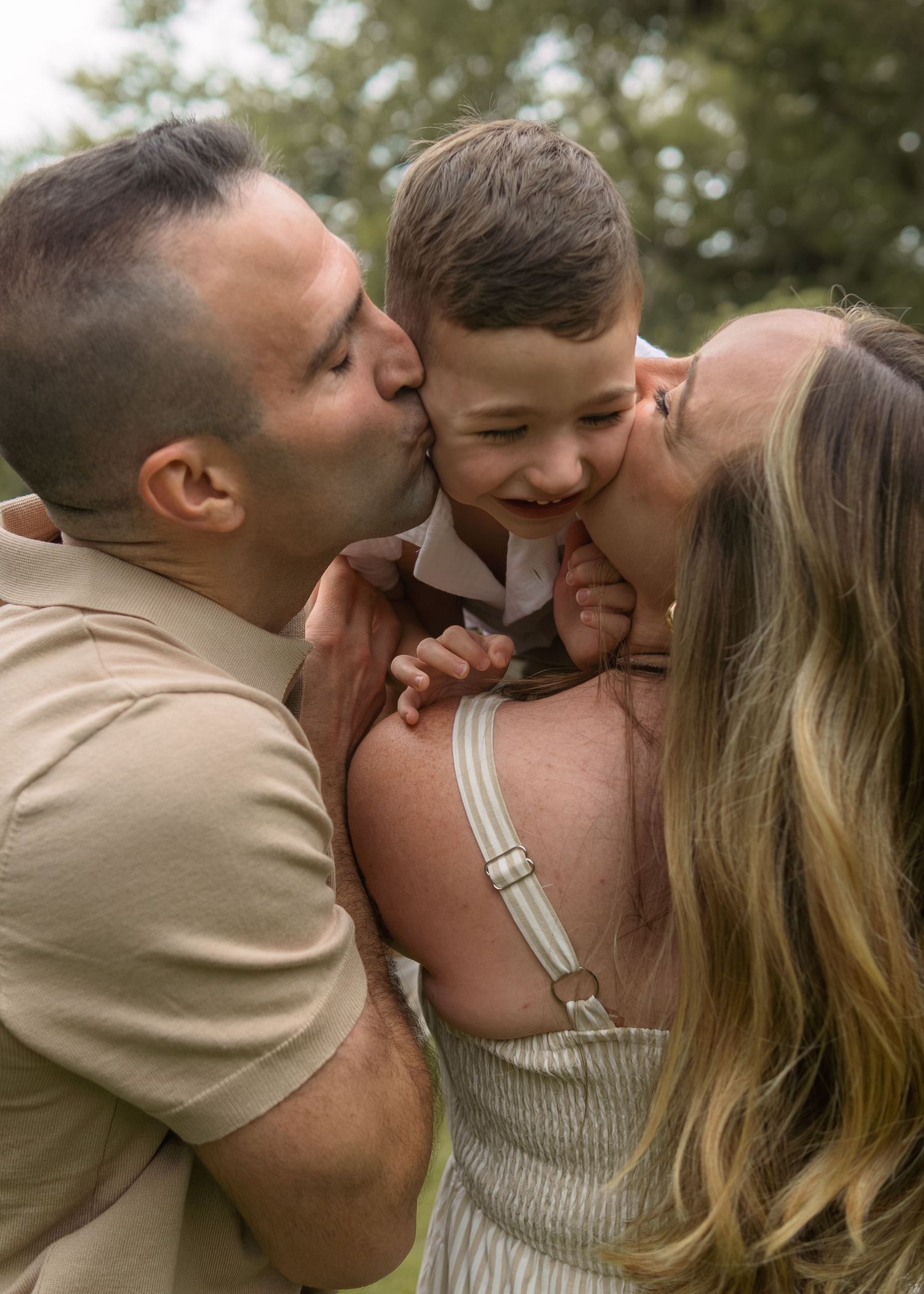 Two parents kissing their young smiling boy on the cheeks outdoors in a loving family portrait.