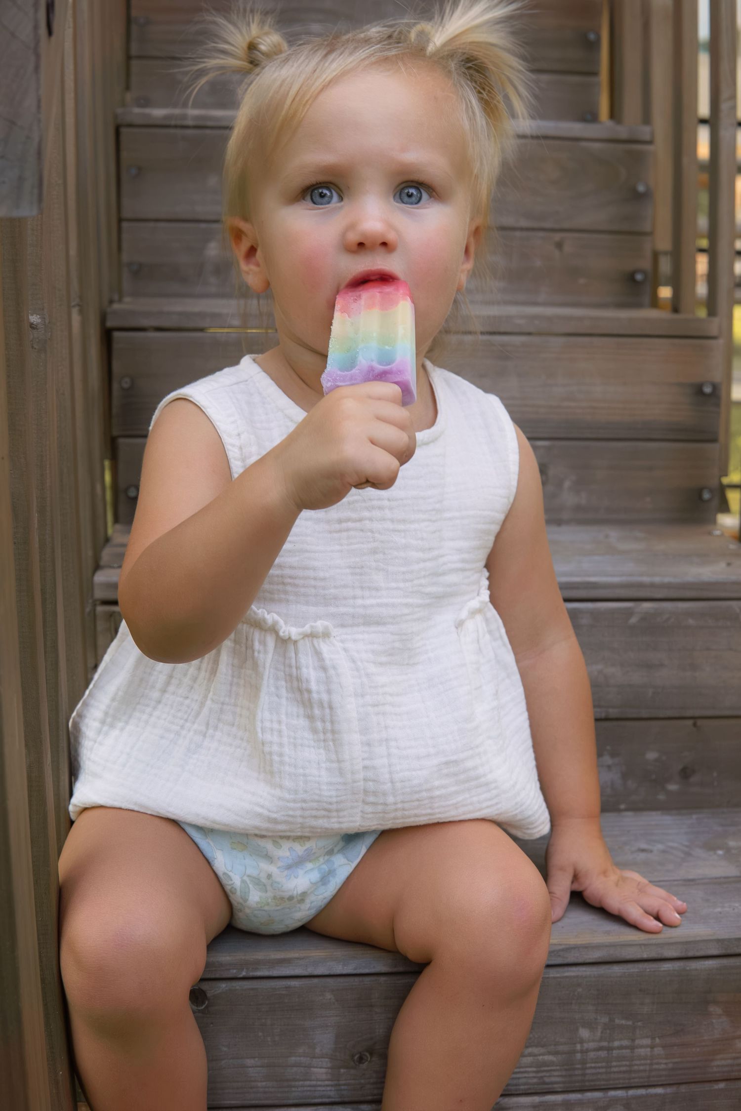 Cute toddler girl with pigtails eating a colorful rainbow popsicle while sitting on wooden steps.