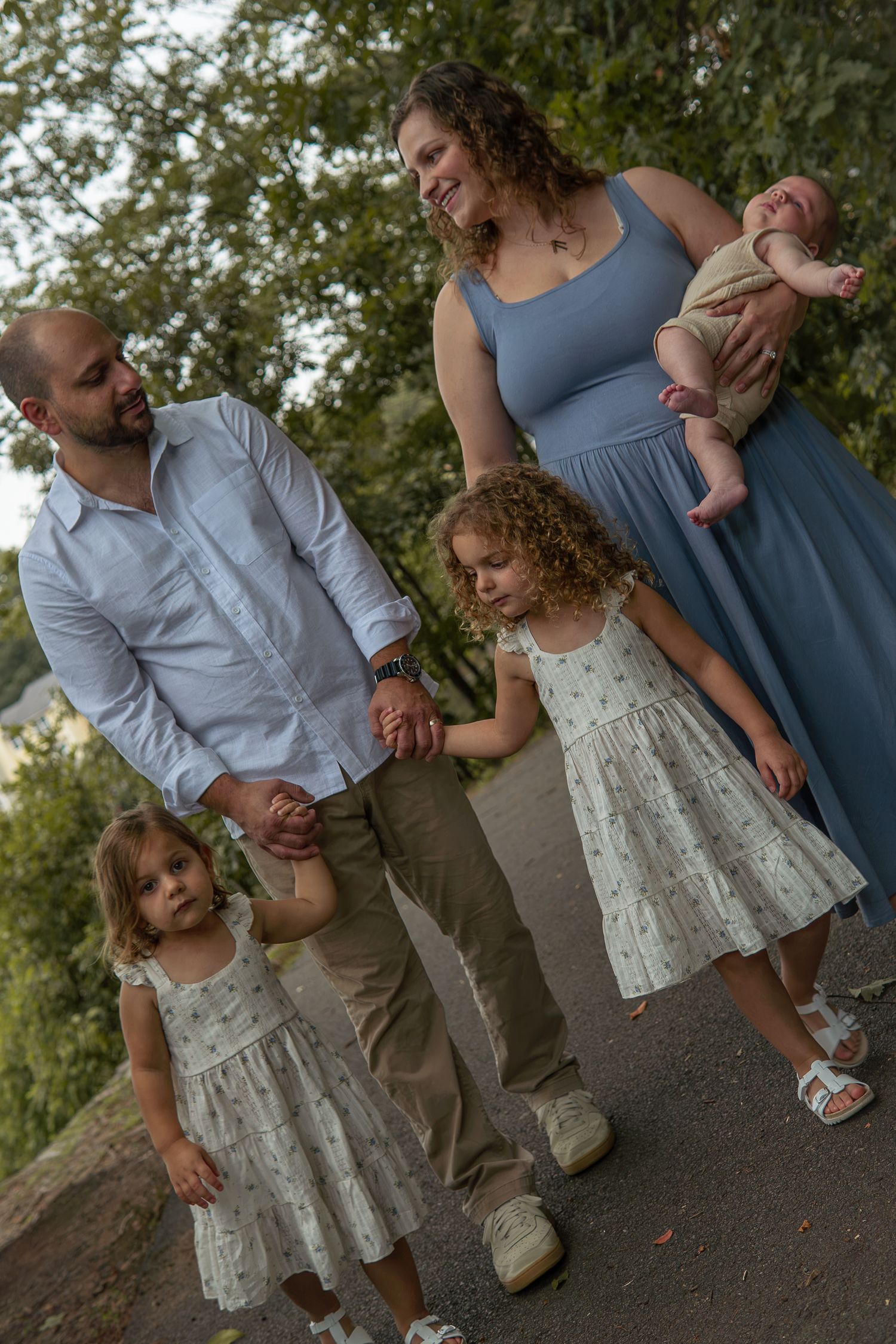 Family of five walking outdoors, parents holding hands with two girls in floral dresses while mom carries newborn baby.