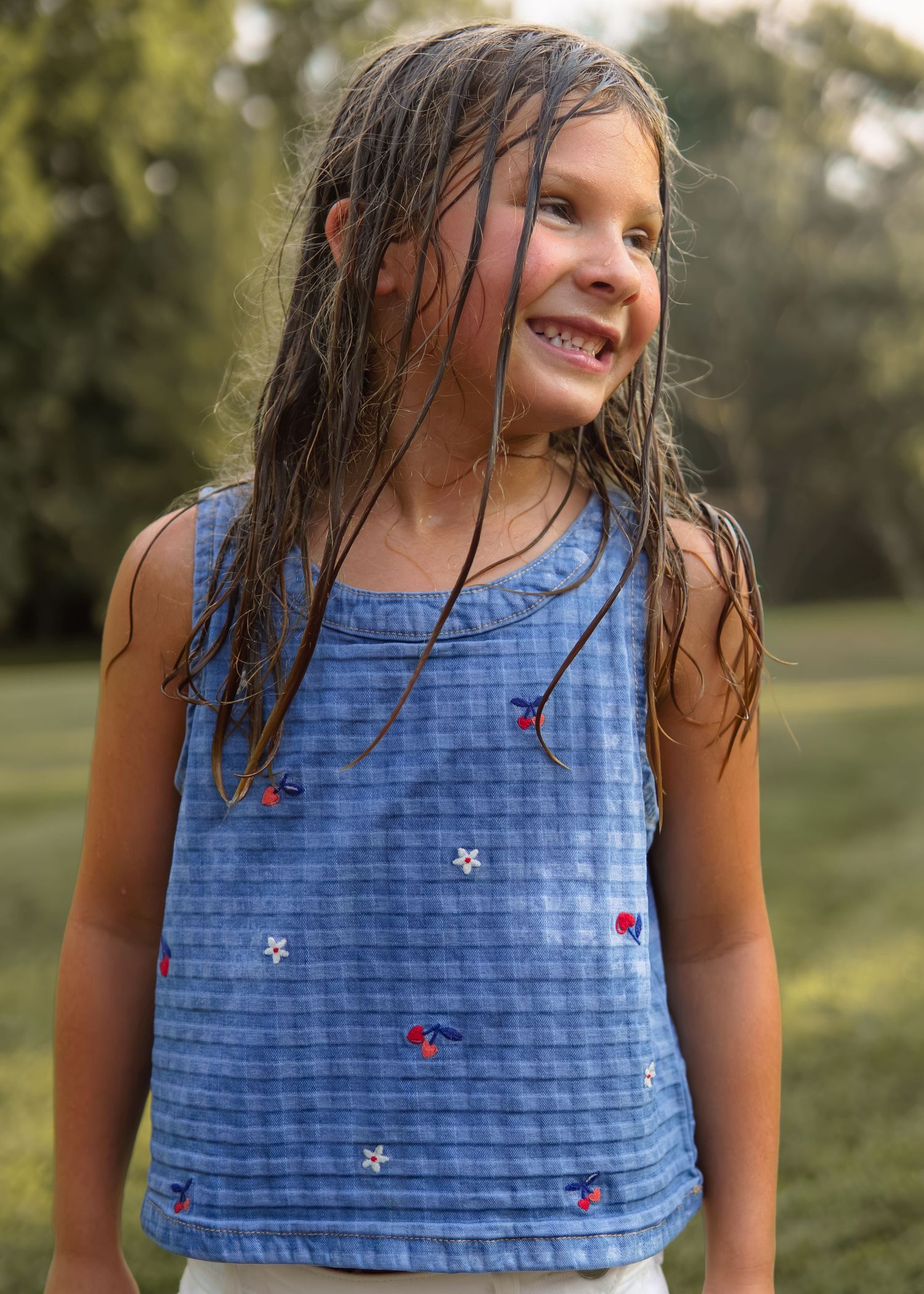 Young girl with wet hair smiling outdoors wearing a blue embroidered sleeveless top in a garden setting.