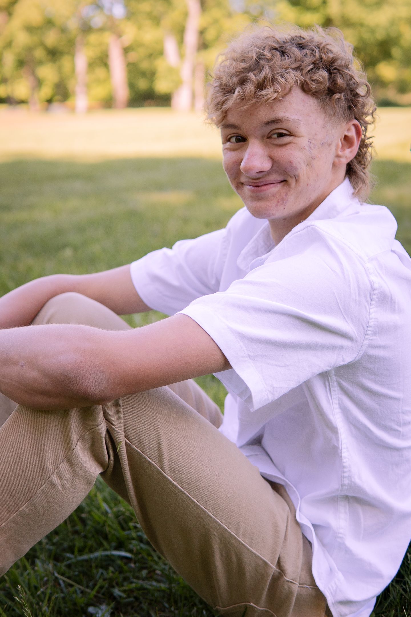 Person in a white button-down shirt and khaki pants sitting on grass on a sunny day.