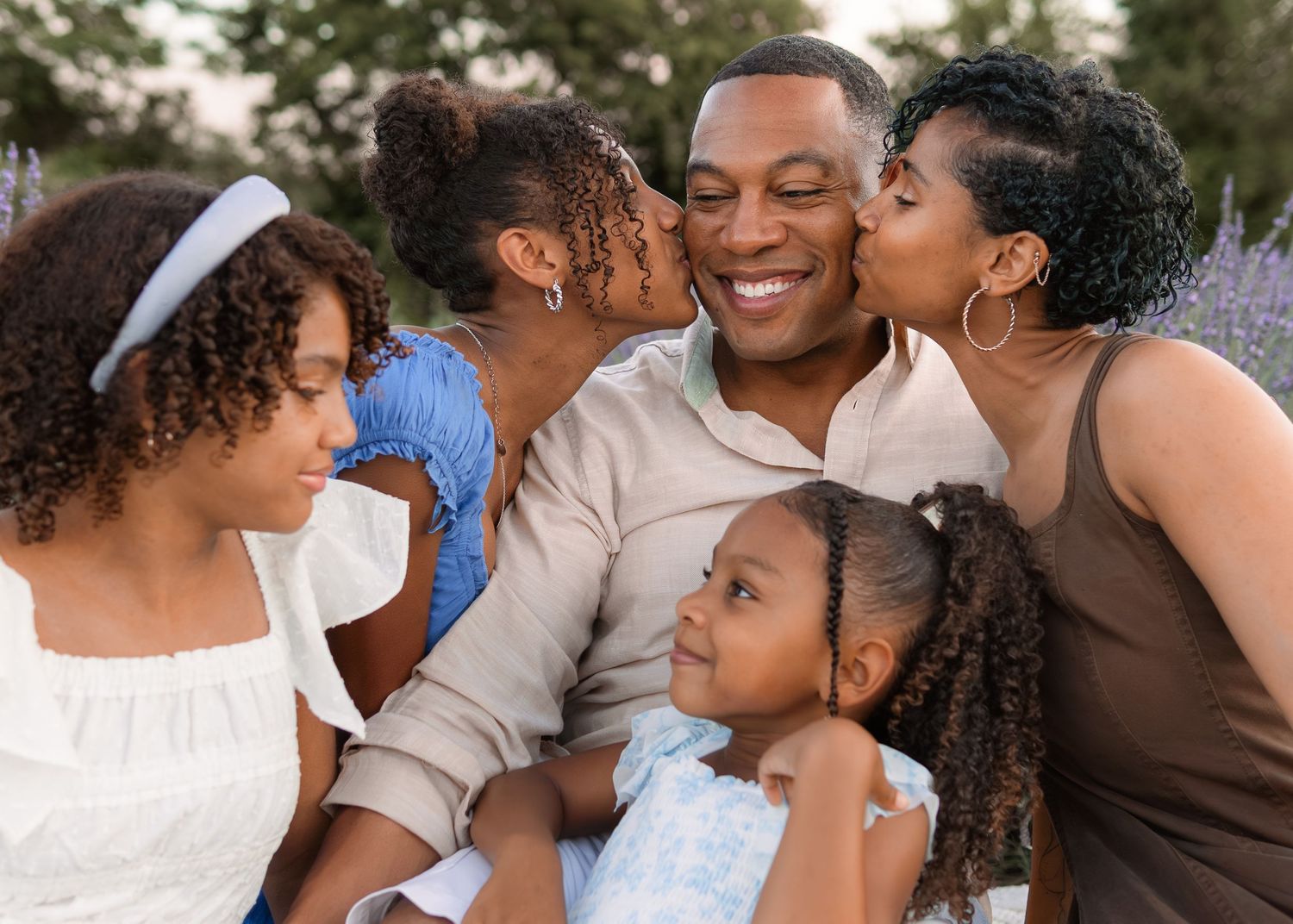 A happy Black family shares a joyful moment outdoors, with children and a woman kissing a smiling man.